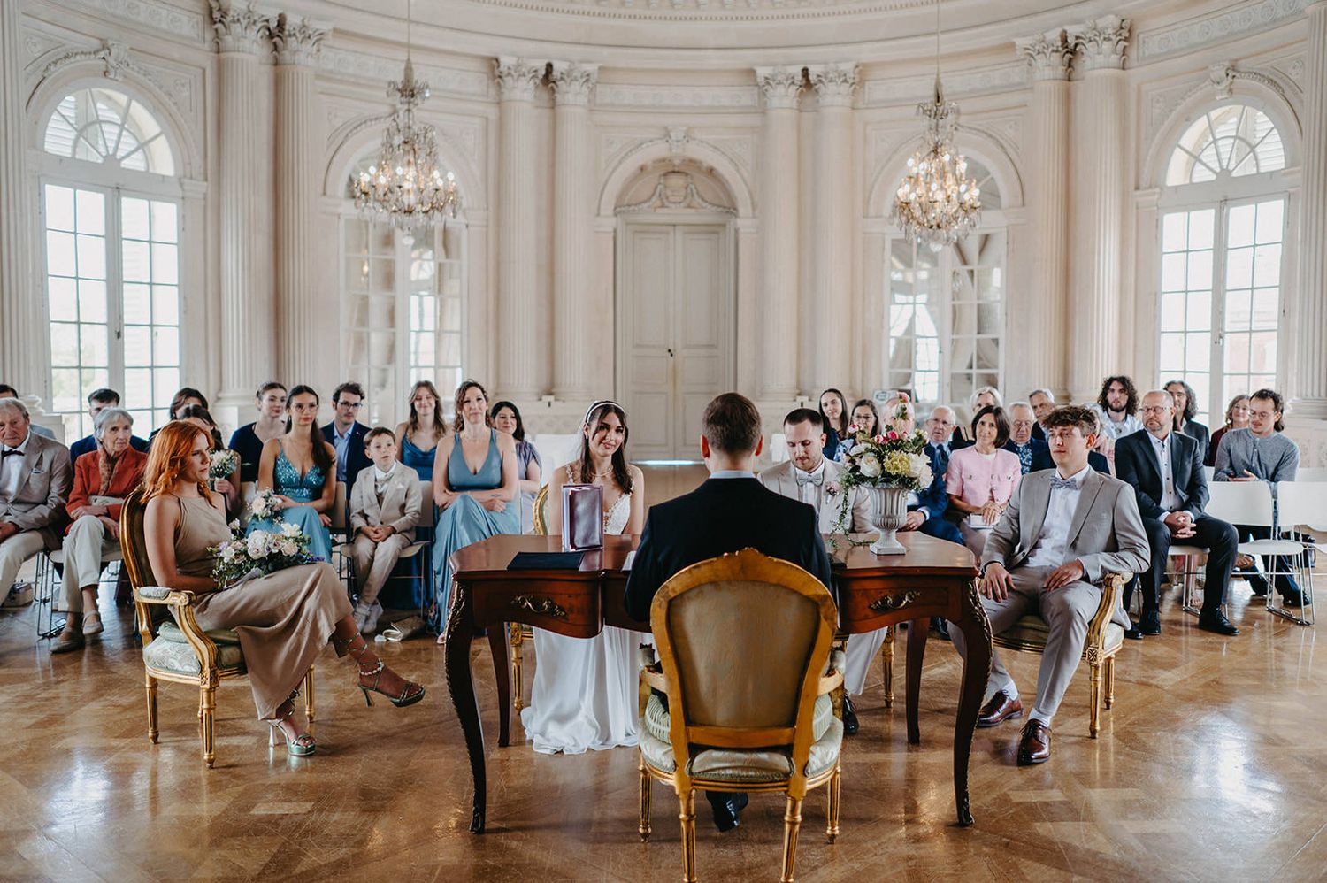 A classical music performance in an elegant ballroom with crystal chandeliers and arched windows as audience members watch.