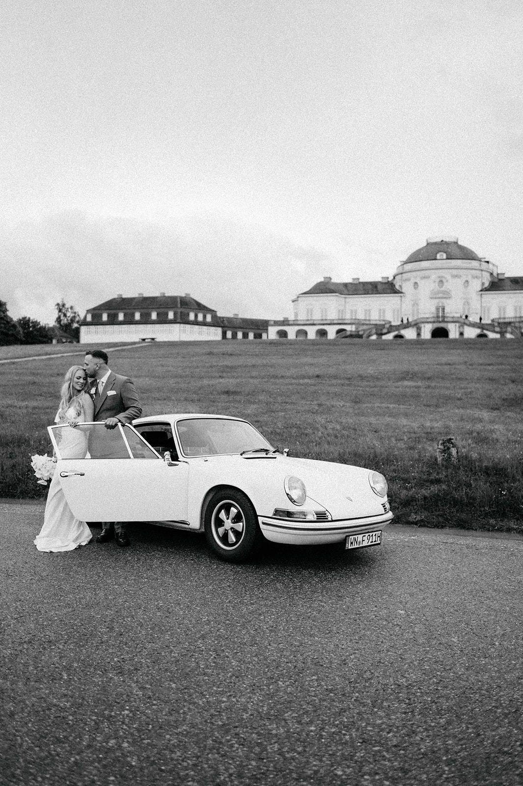 A vintage wedding scene with a classic convertible Porsche 911 parked in a grassy field near buildings.