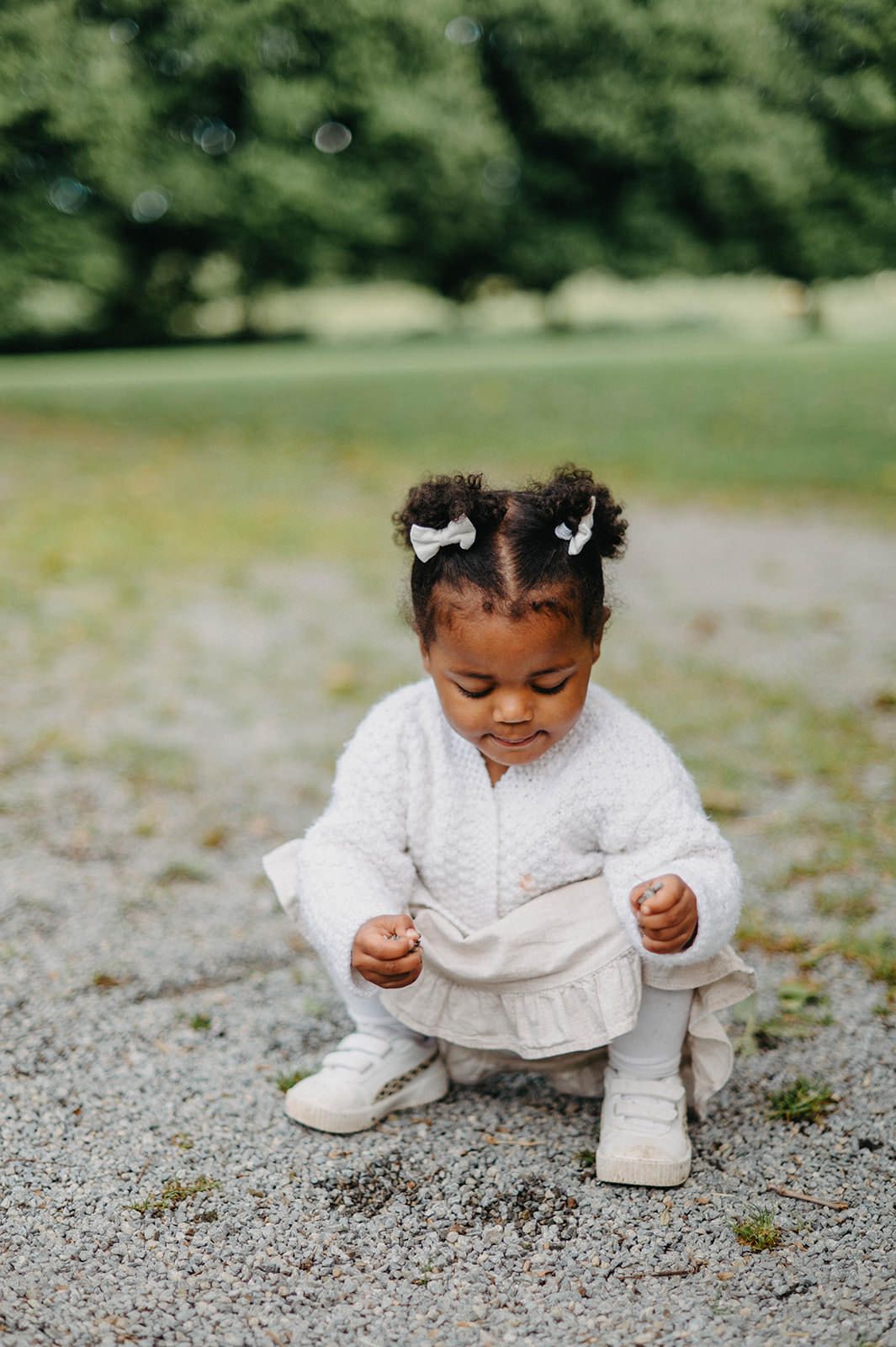 A small child in a white dress sits on gravel while looking down and playing with something on the ground.