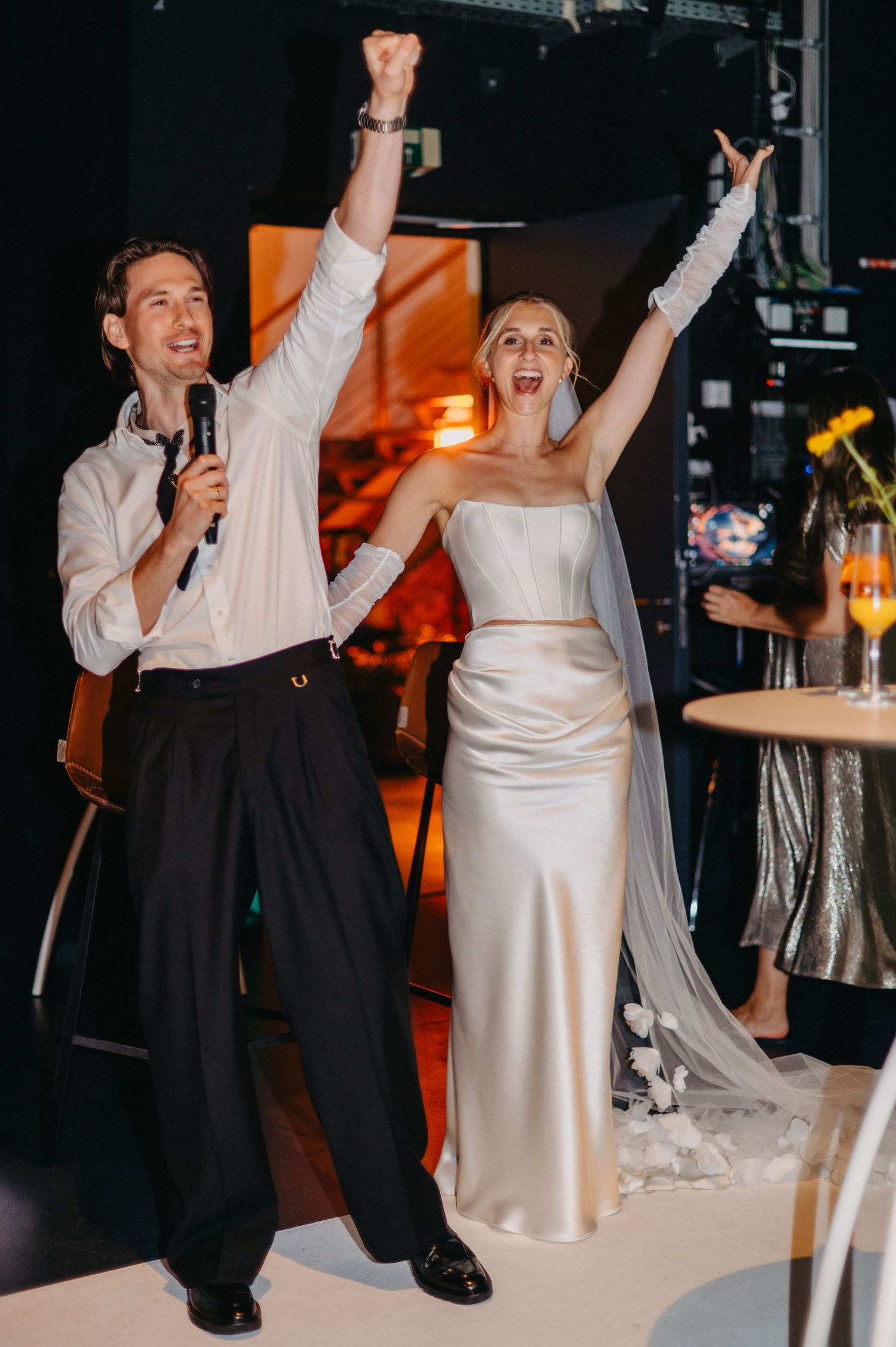 Two people in formal attire dance and celebrate joyfully at a wedding reception with warm lighting in the background.