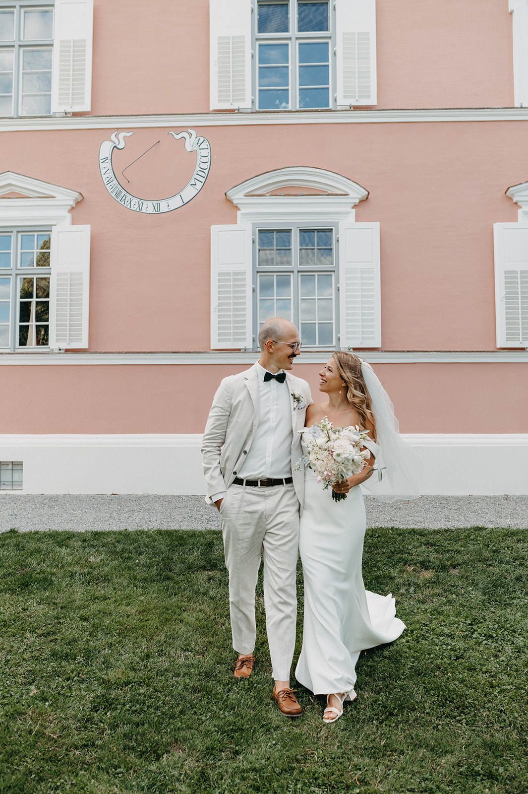A couple walks along the grass in front of a pastel pink historic building with classical architecture.