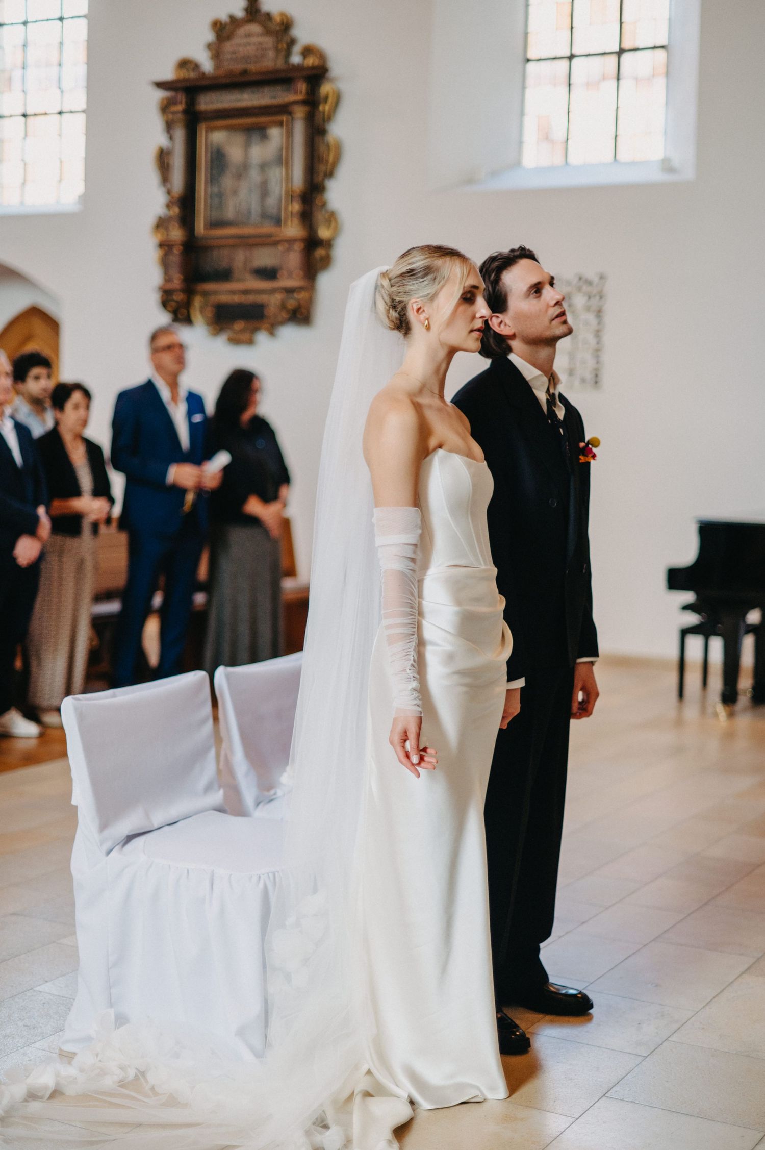 A bride and groom stand together in a bright white church interior during their wedding ceremony.