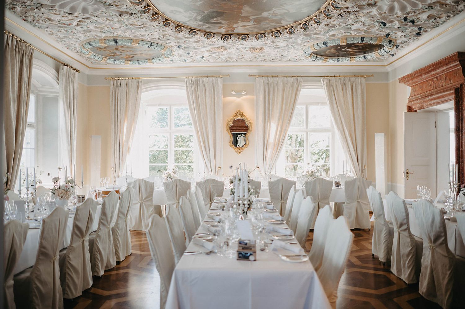 Elegant white wedding reception hall with ornate ceiling details, draped chairs, and natural light streaming through windows.