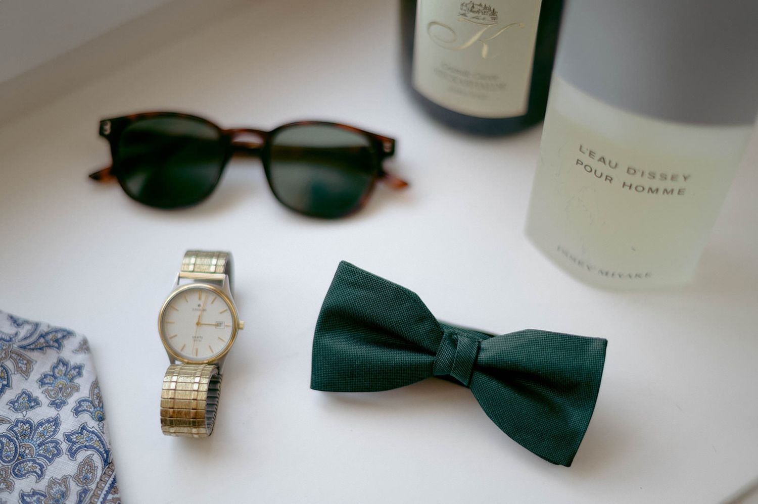 Wedding accessories including a black bow tie, sunglasses, and wristwatch arranged on a white surface.