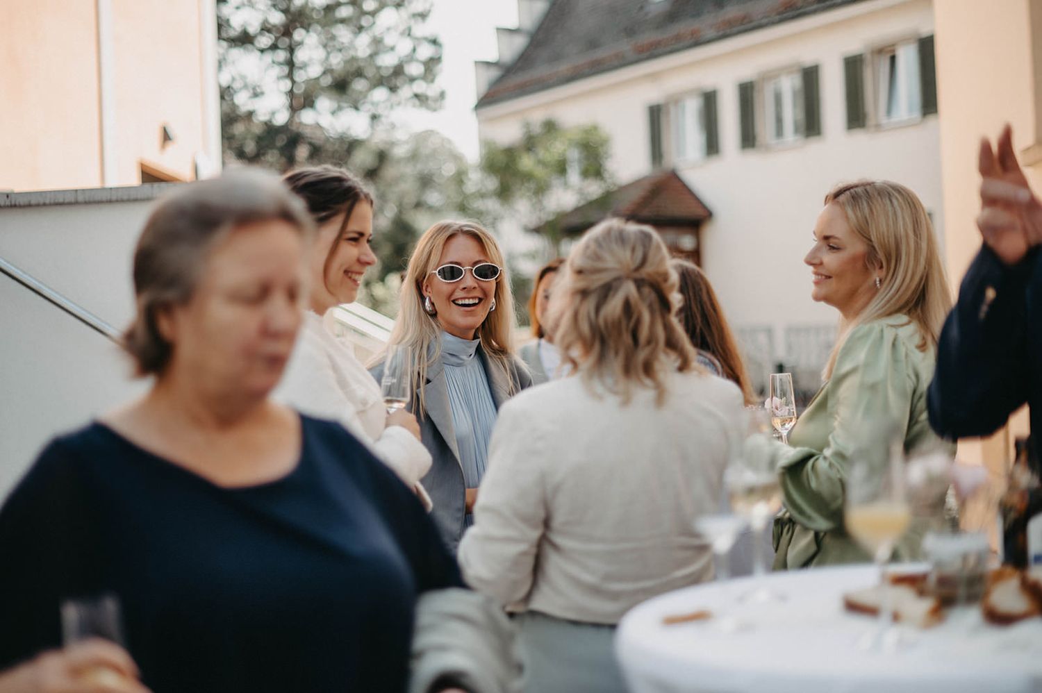 Guests gathered outdoors at a social event share laughter and conversation around tables during a celebration.