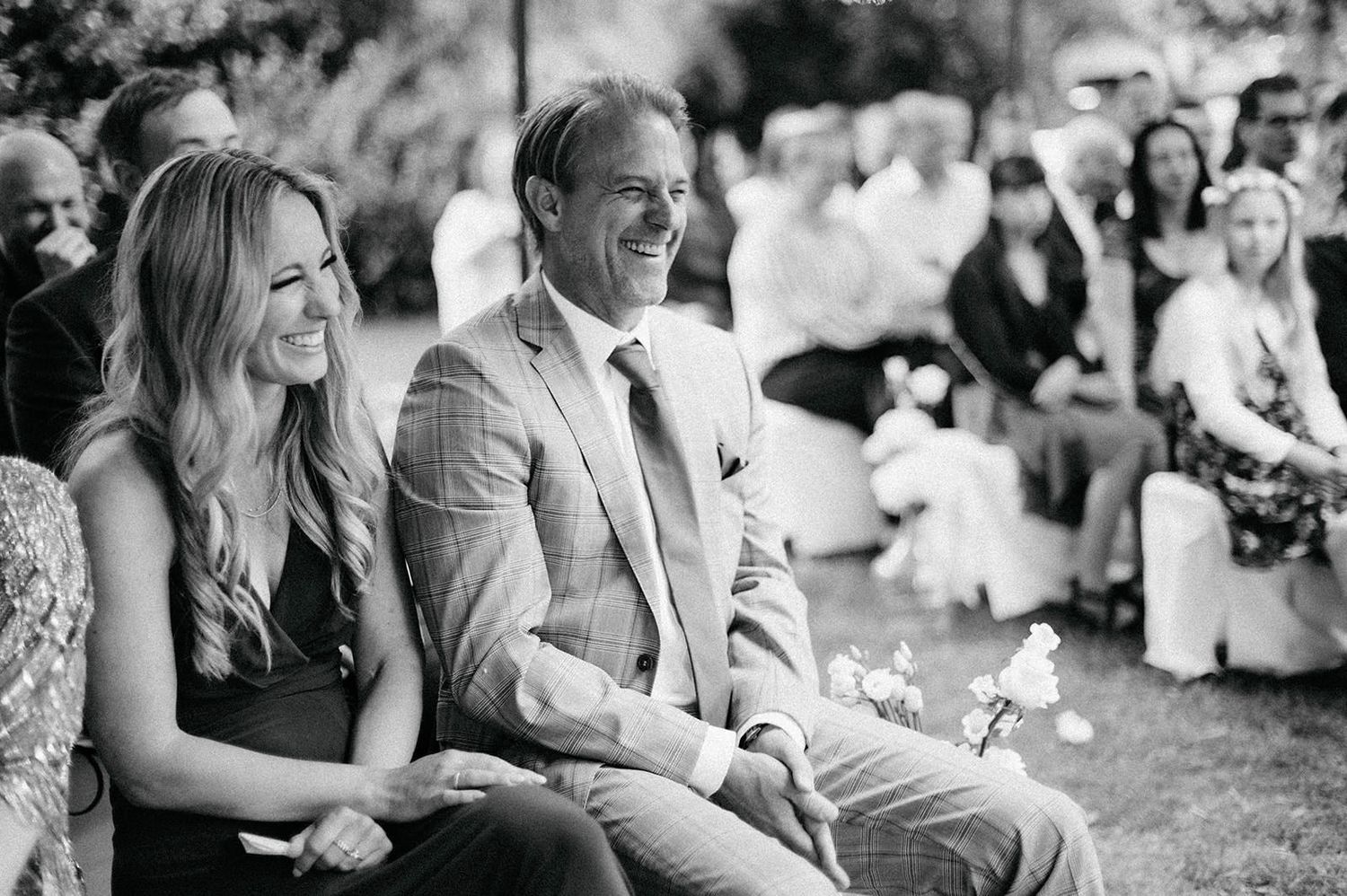 Wedding guests sit outdoors on white chairs during a sunny garden ceremony.