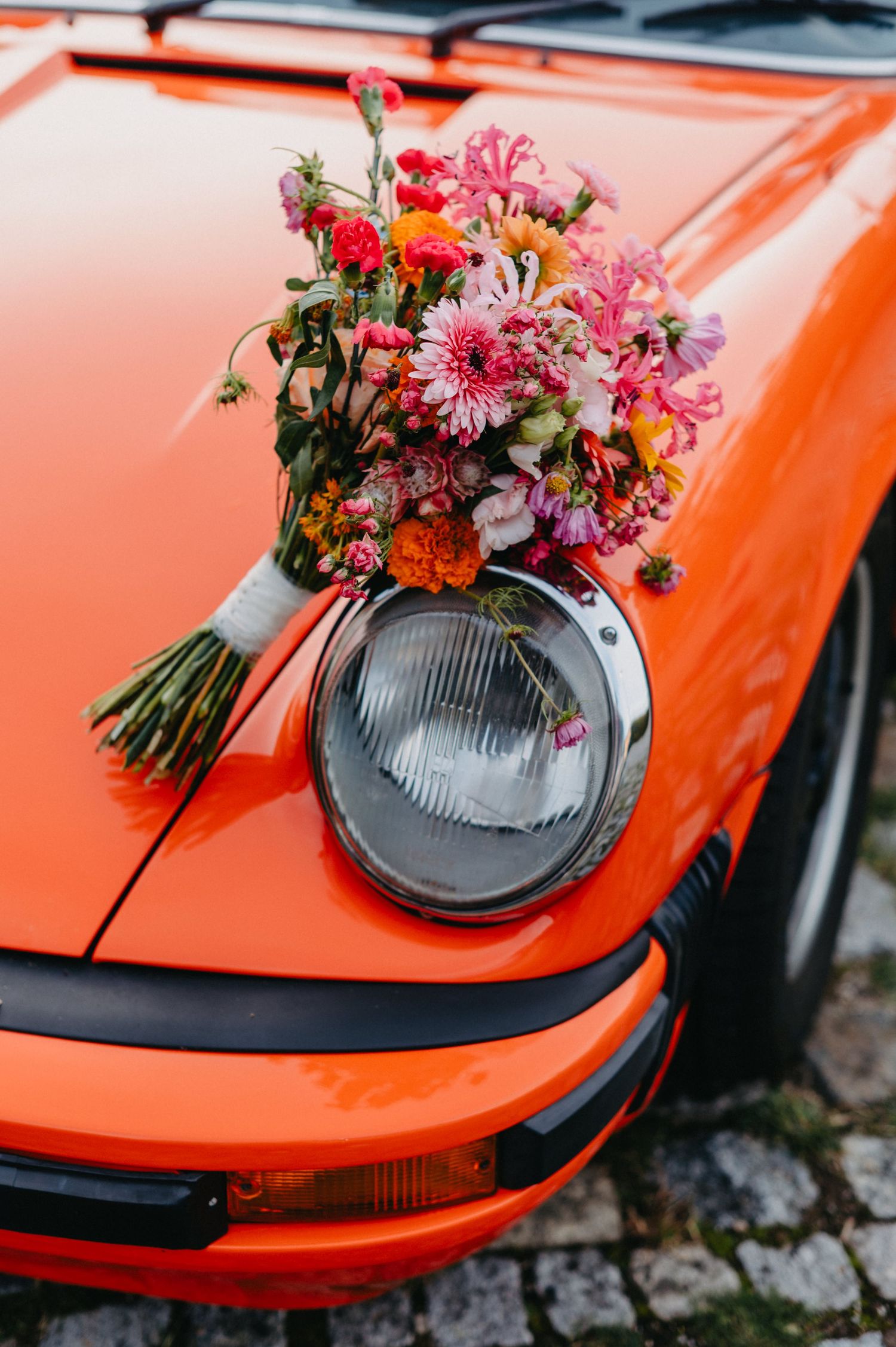 A vibrant pink and red flower bouquet rests on the headlight of an orange vintage Porsche car.