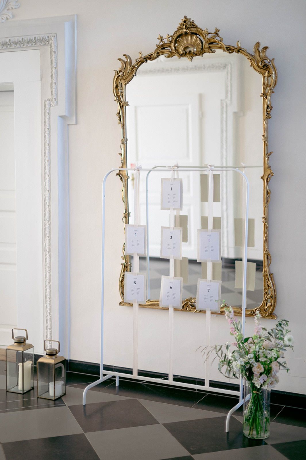 Elegant ornate gold-framed mirrors hang on white walls above dark counters with delicate floral arrangements.