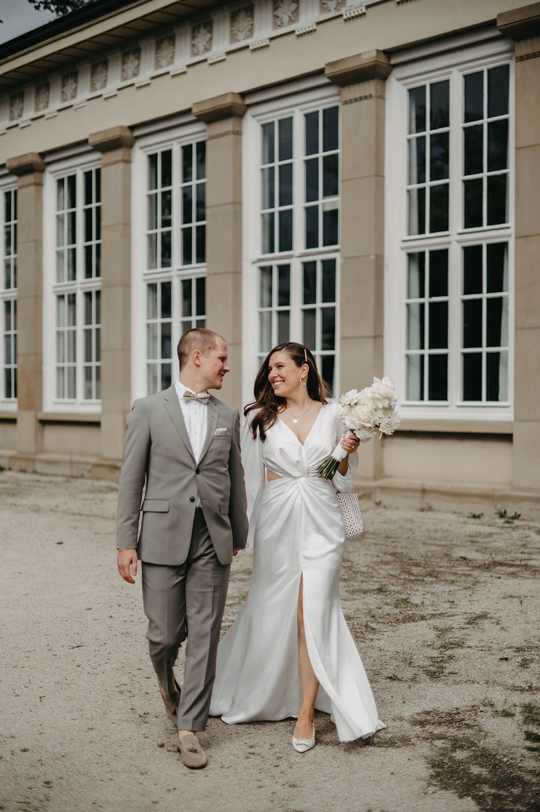 Newlyweds walking together and smiling outside historic building with large white windows.