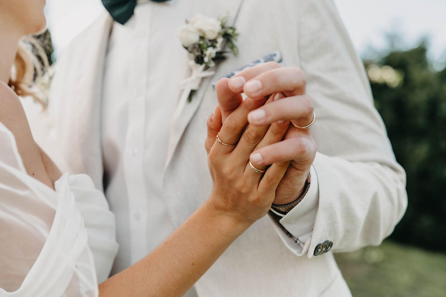 Close-up shot of newlywed couple holding hands during outdoor wedding ceremony, with white suit sleeve visible.