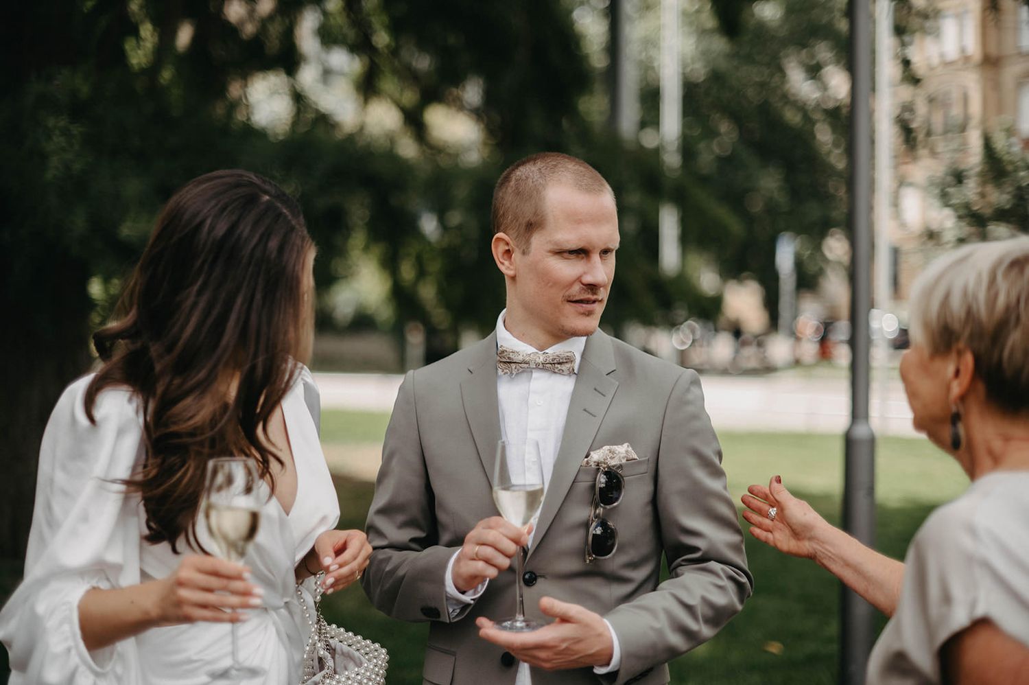 Three people engaged in casual conversation while holding drinks at an outdoor gathering in a park setting.