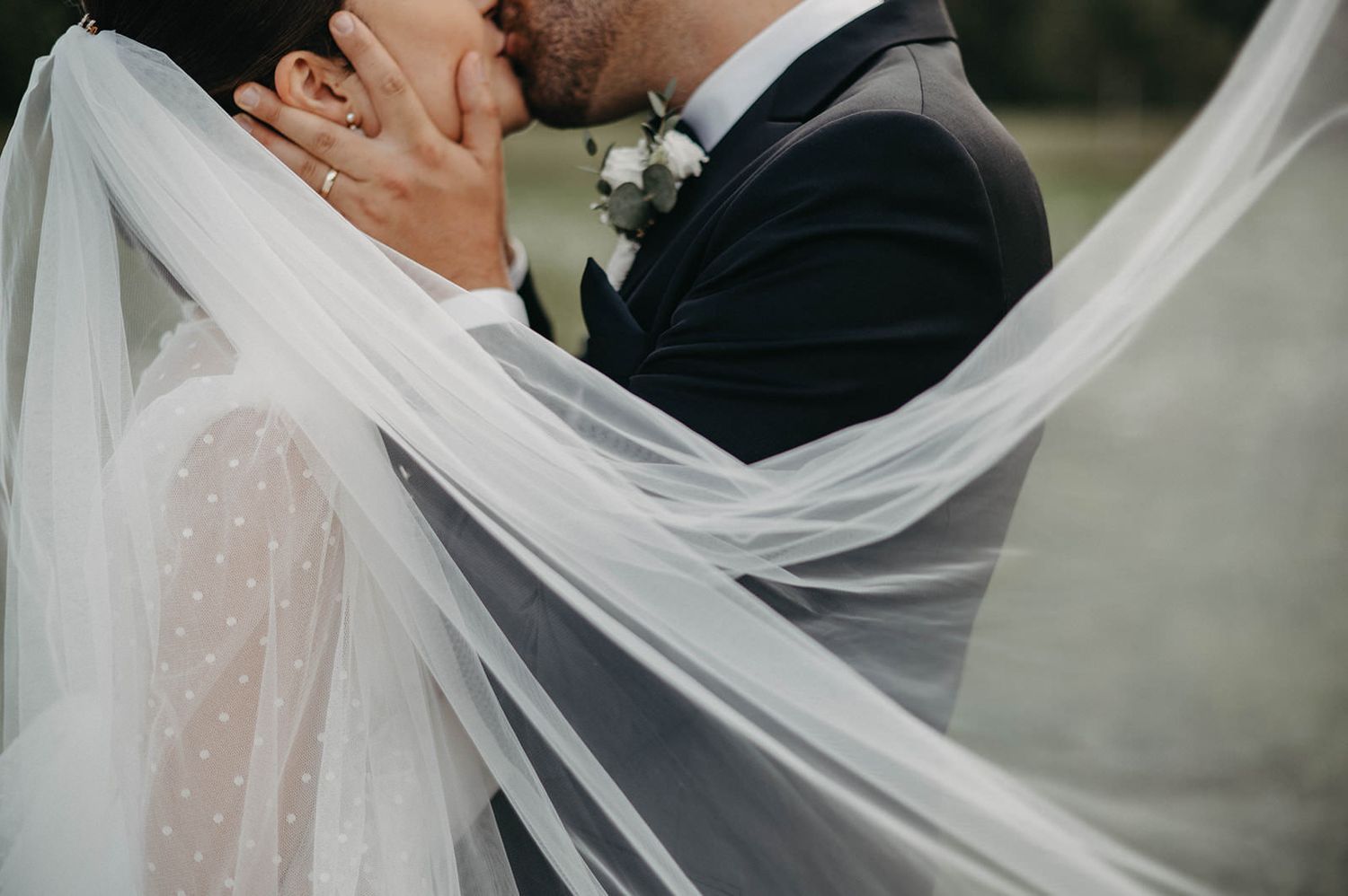 A bride and groom embrace under a flowing white veil during their outdoor wedding ceremony.
