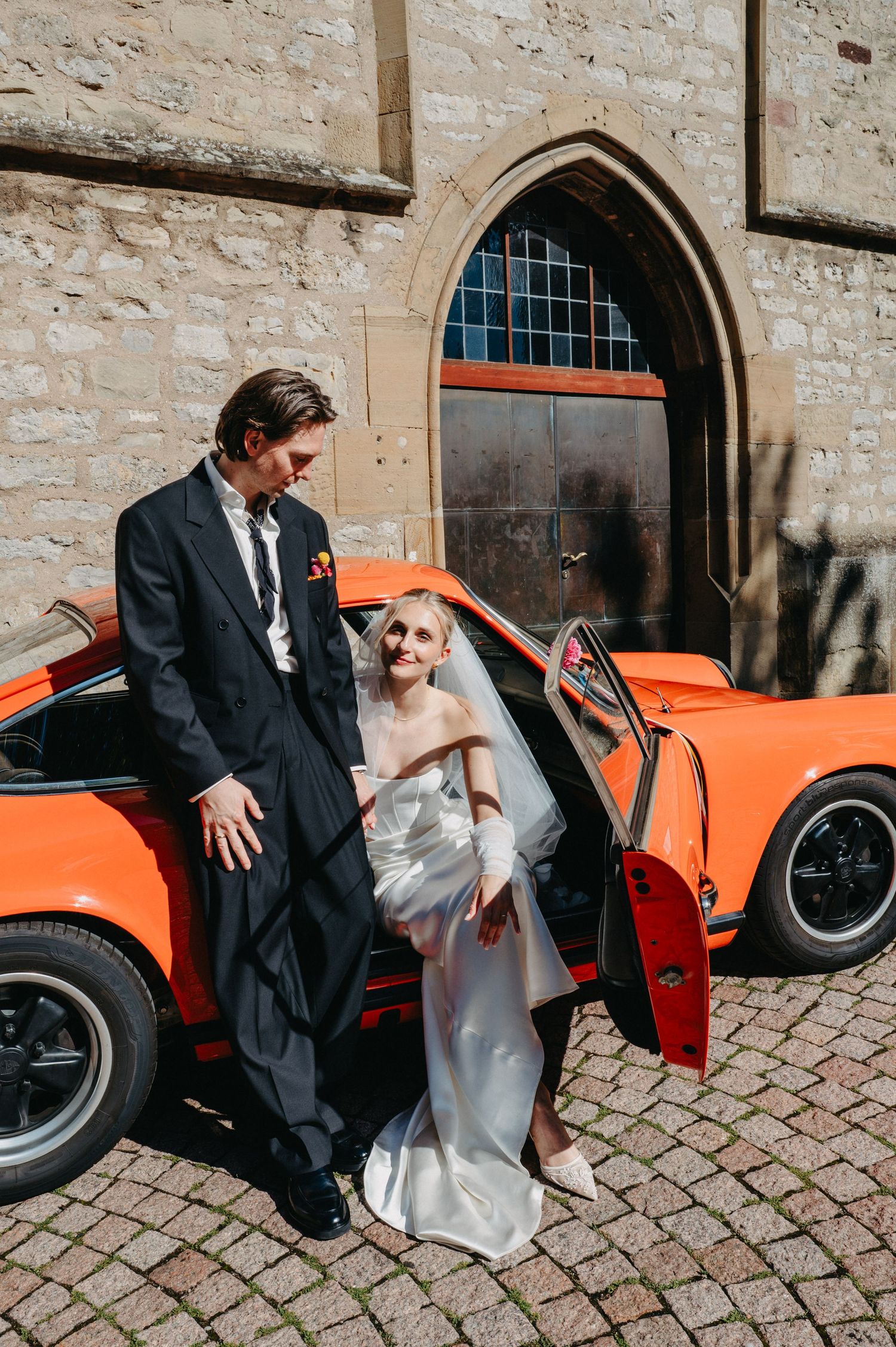 A sequence of wedding photos showing a couple getting into a bright orange vintage sports car outside a stone building.