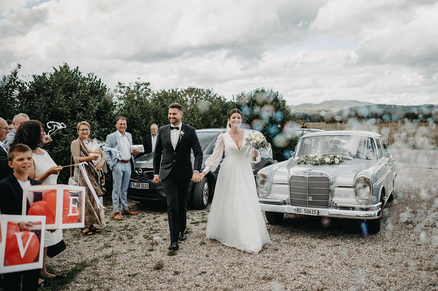 A bride and groom walk hand in hand past guests and a vintage Mercedes on a rural hilltop during their wedding celebration.
