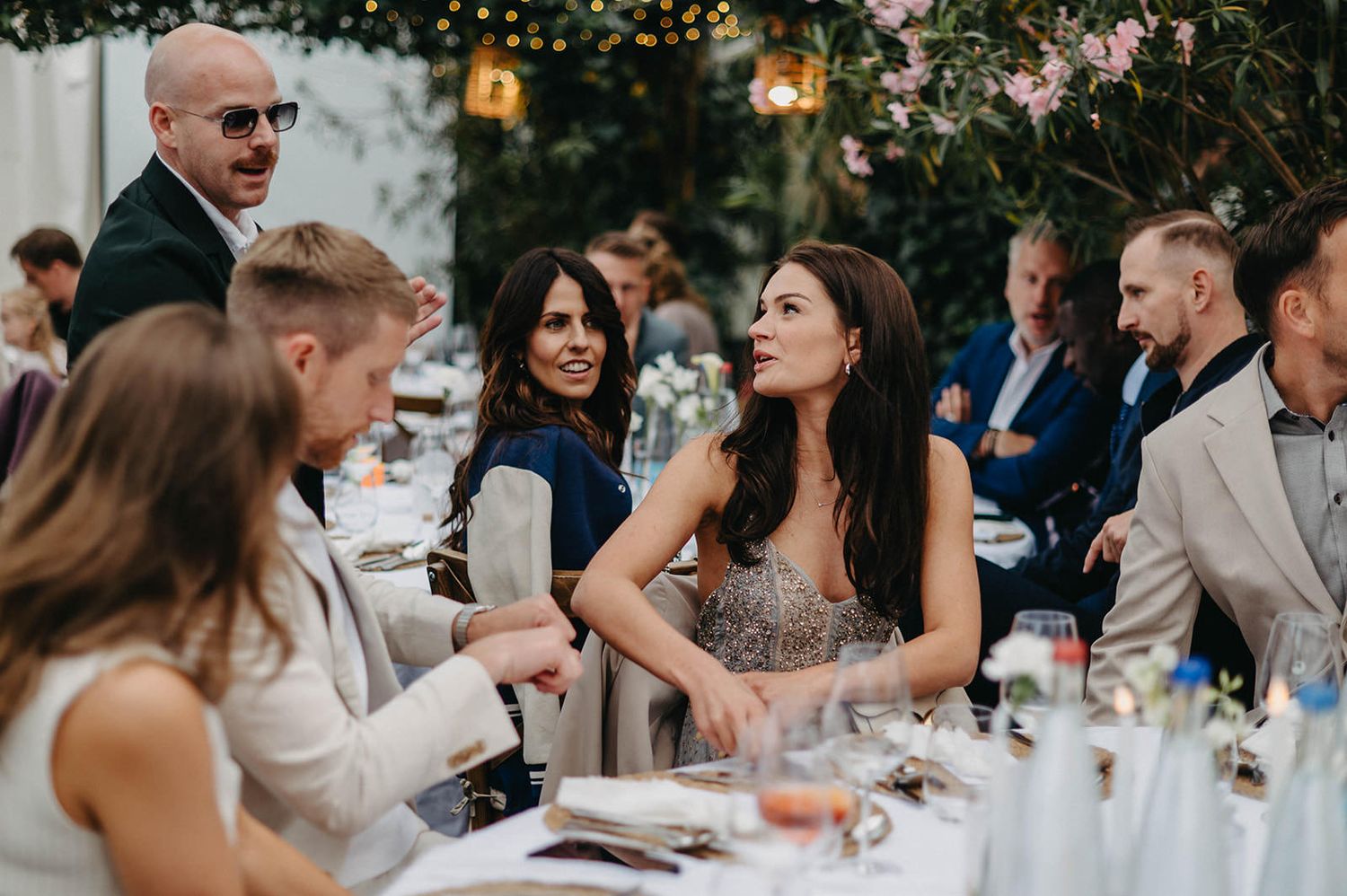 Wedding guests enjoy an outdoor reception dinner under string lights and greenery at dusk.