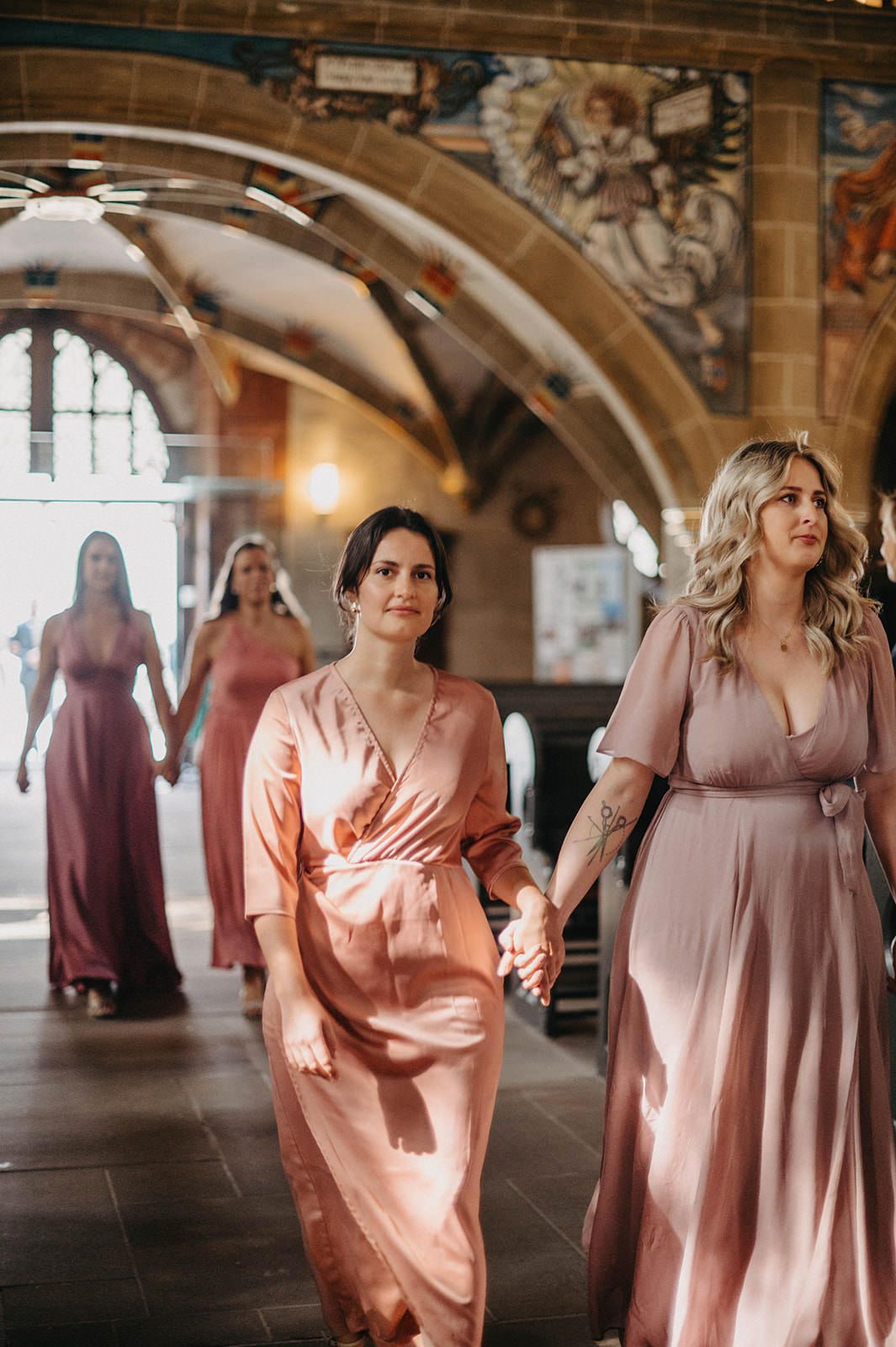 Bridesmaids in flowing pink and mauve dresses walk through an elegant arched hallway with ornate architectural details.