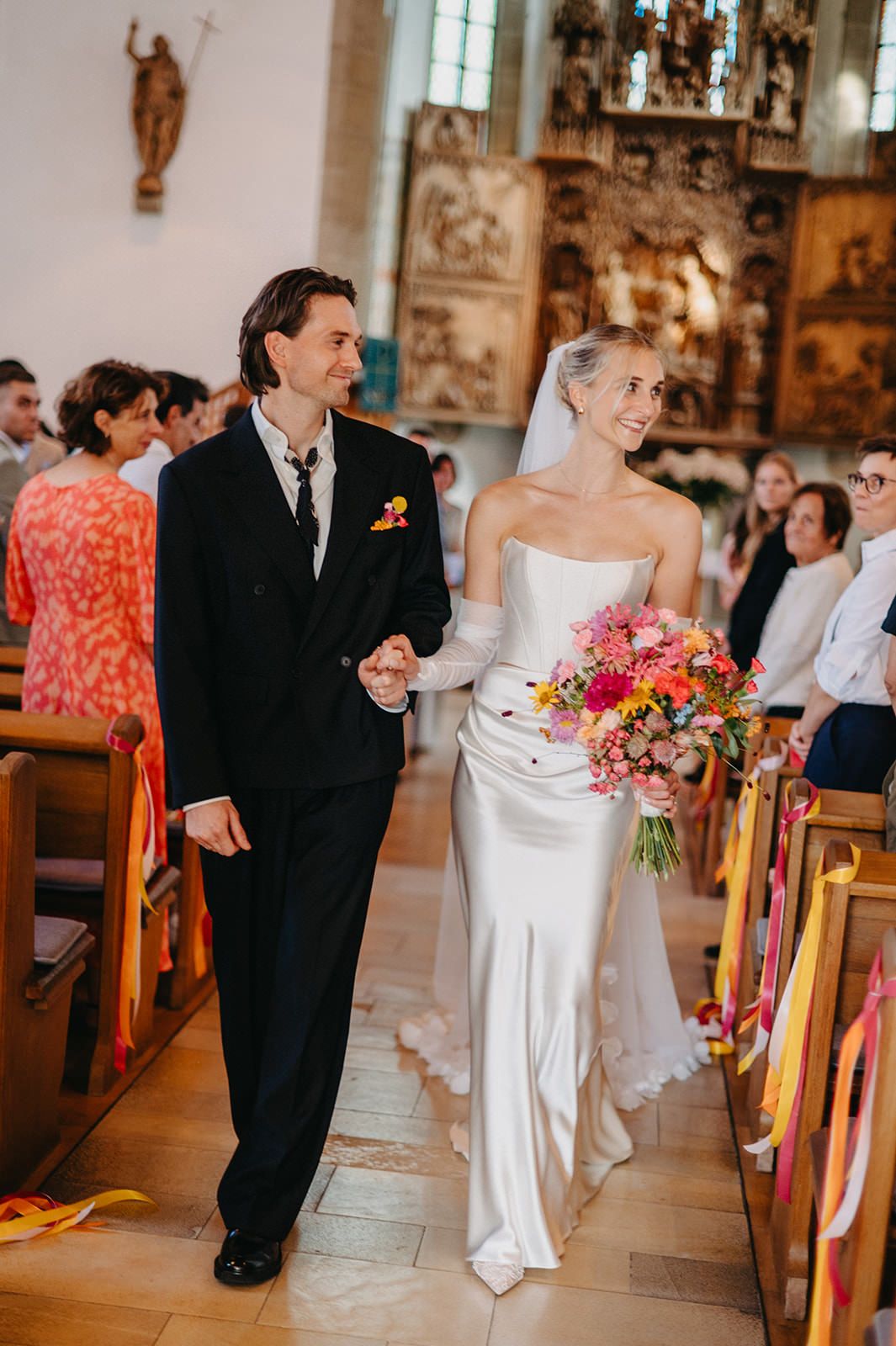 A couple walks down the church aisle during their wedding ceremony, with colorful floral decorations and guests in view.
