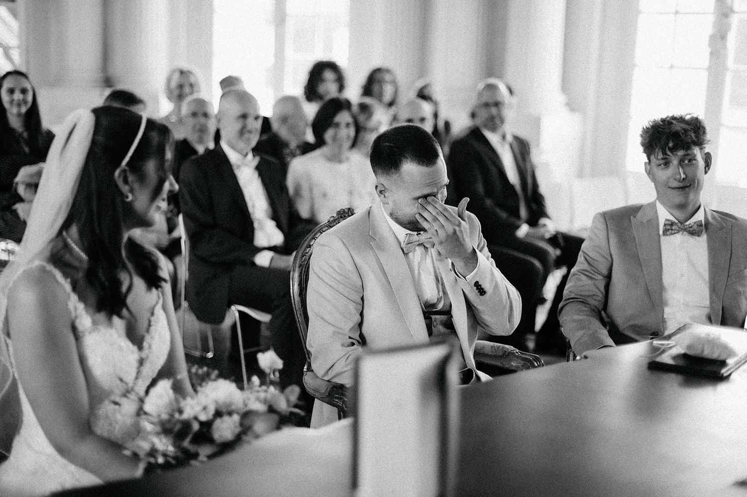 Black and white photo of wedding guests seated at tables during a reception in a bright indoor space.
