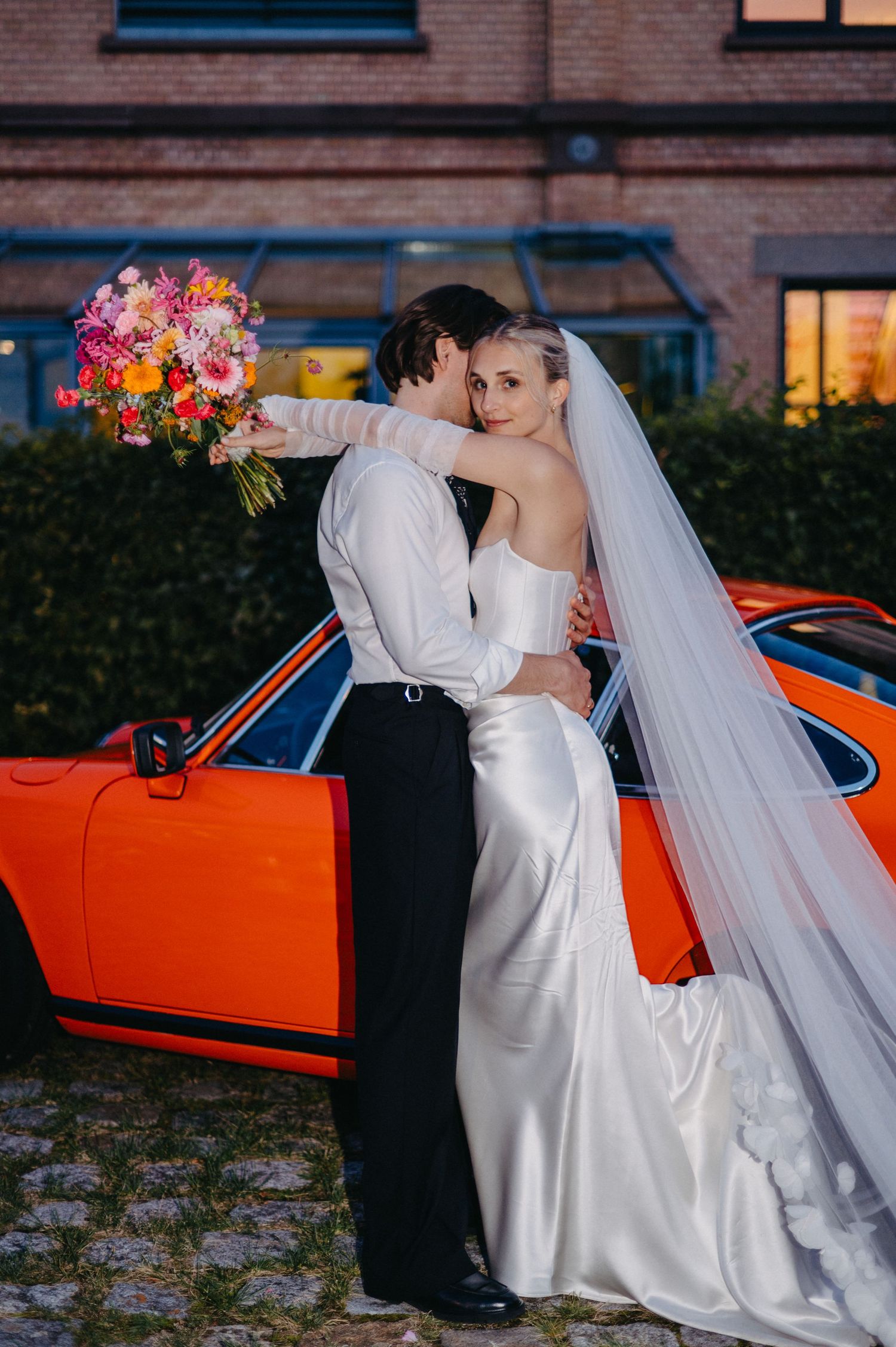 A romantic wedding couple embraces beside a bright orange vintage car while holding a vibrant floral bouquet.