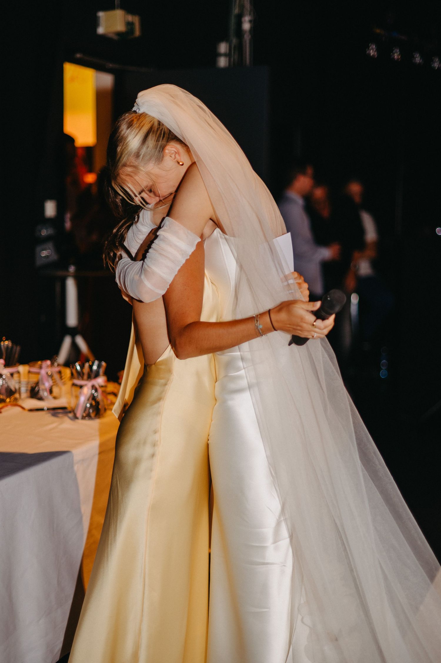 Two figures in formal wedding attire share an intimate embrace during a wedding reception.