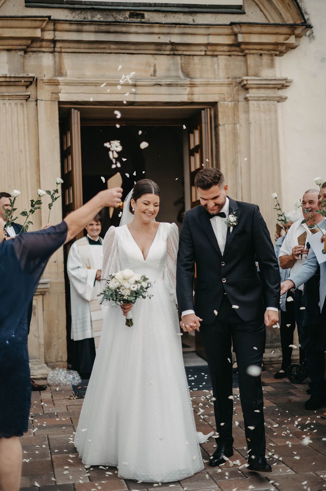 Wedding guests throw white flower petals as newlyweds exit stone building in celebration.