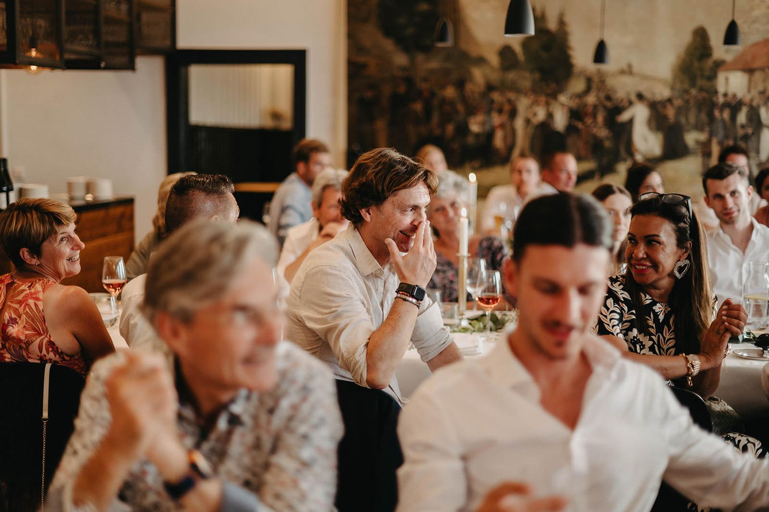 Wedding guests laugh and socialize during an indoor reception at a rustic venue with warm lighting and wooden details.