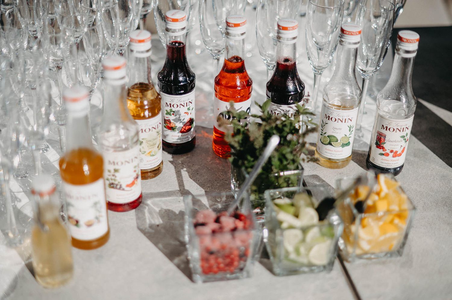Glass bottles of cocktail mixers and garnishes arranged on a bar counter with glassware for drink service.