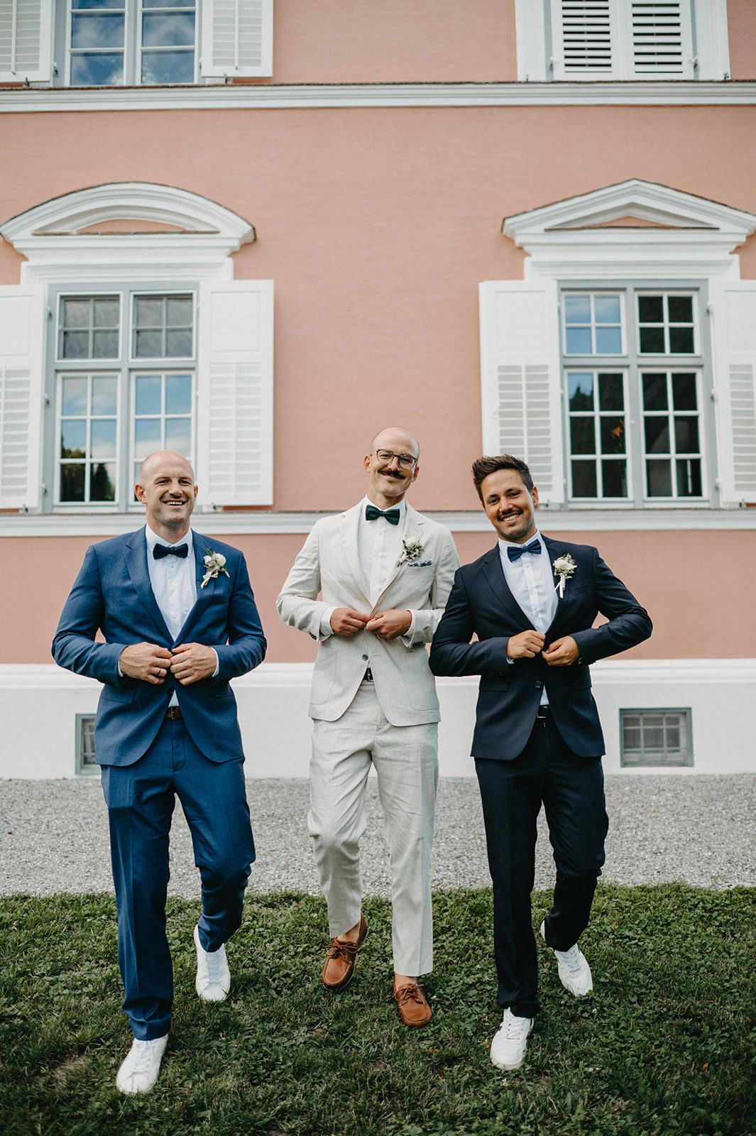 Groomsmen in navy and light colored suits pose playfully on grass in front of pink building with white shutters.