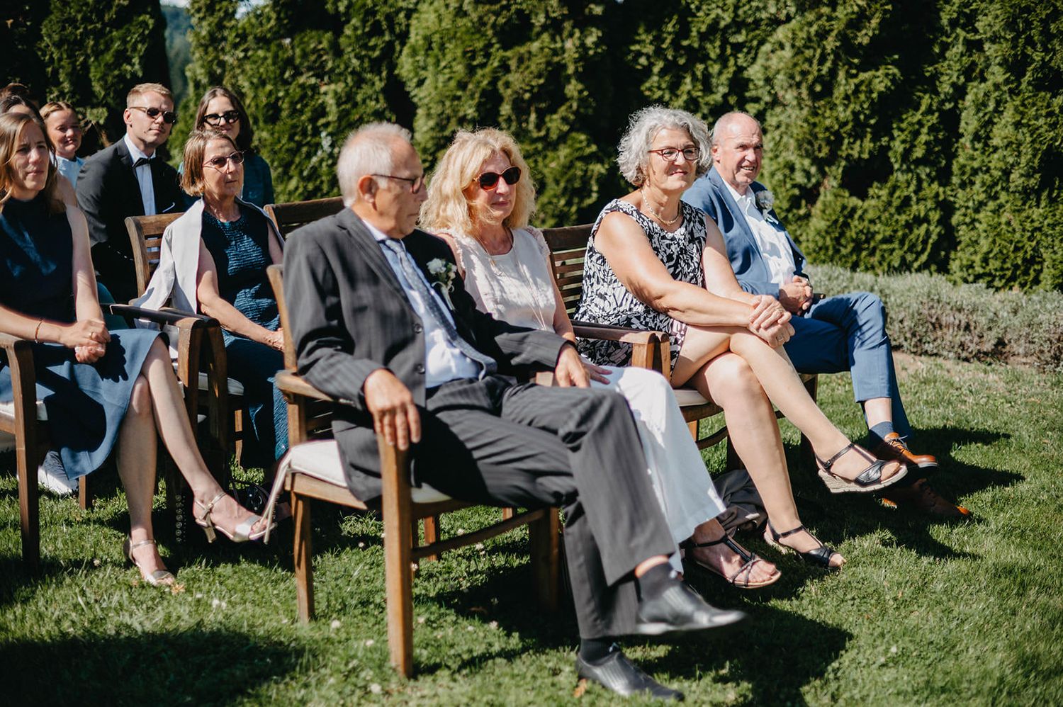 Wedding guests sit on white chairs arranged outdoors on a lush green lawn during a garden ceremony.