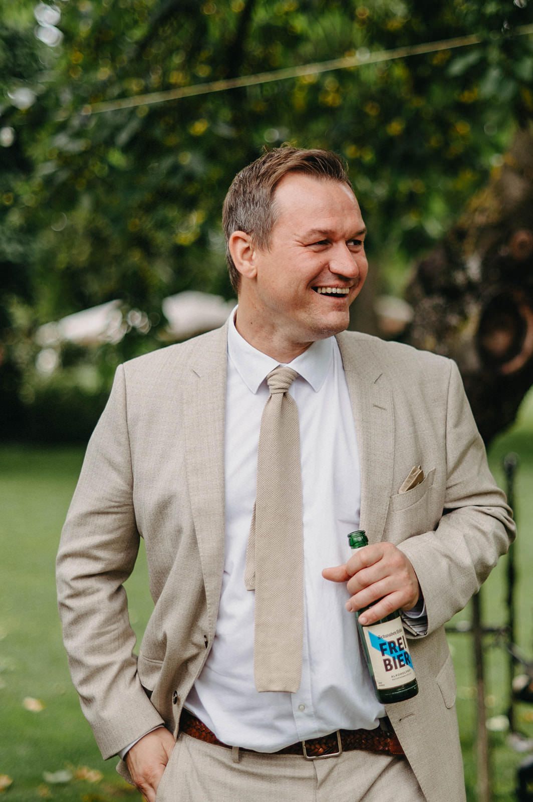 A person in a beige suit and tie smiles while holding a beverage bottle at an outdoor event.