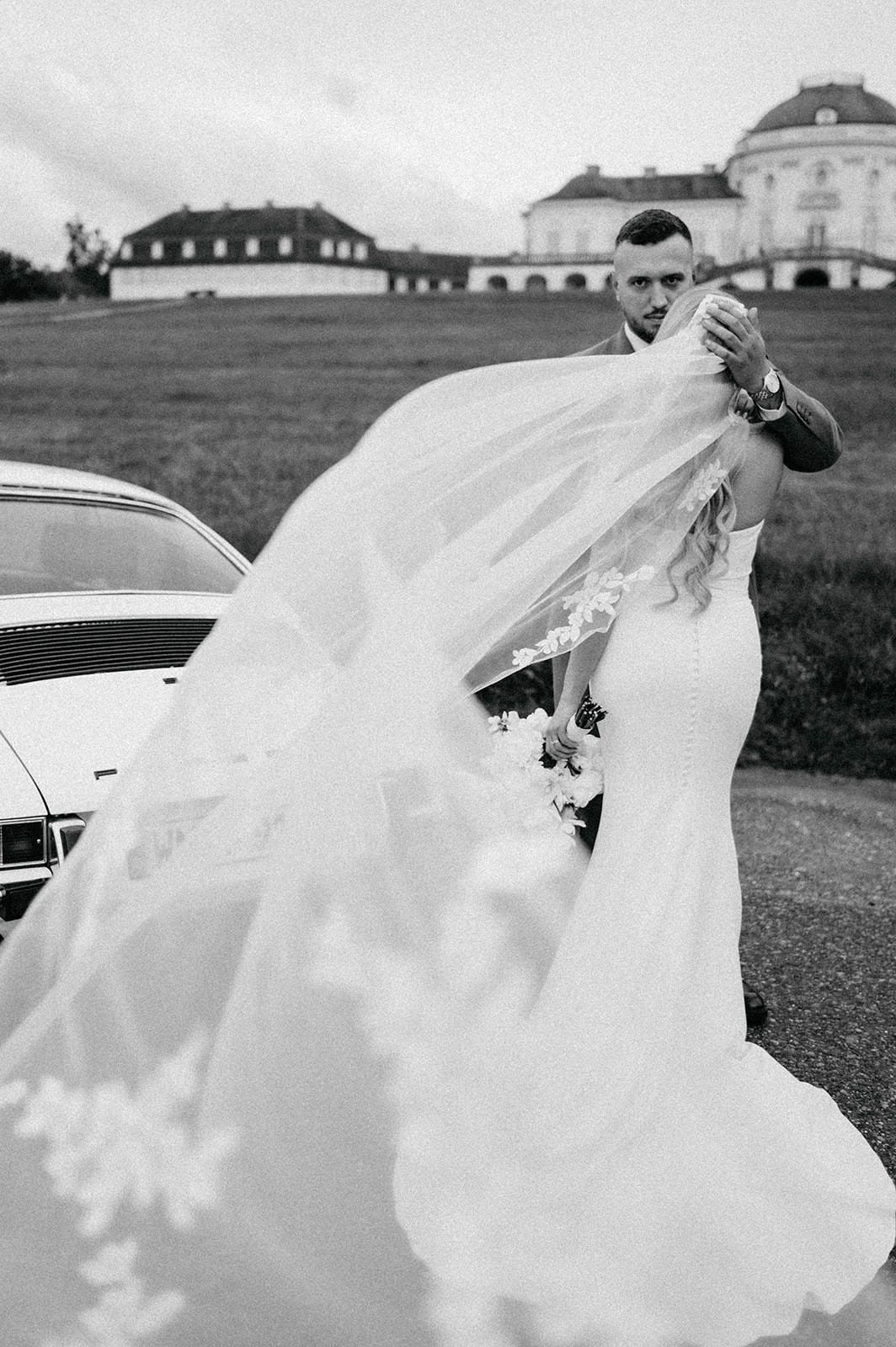 A flowing white wedding veil billows dramatically in the wind against a backdrop of historic buildings on a cloudy day.