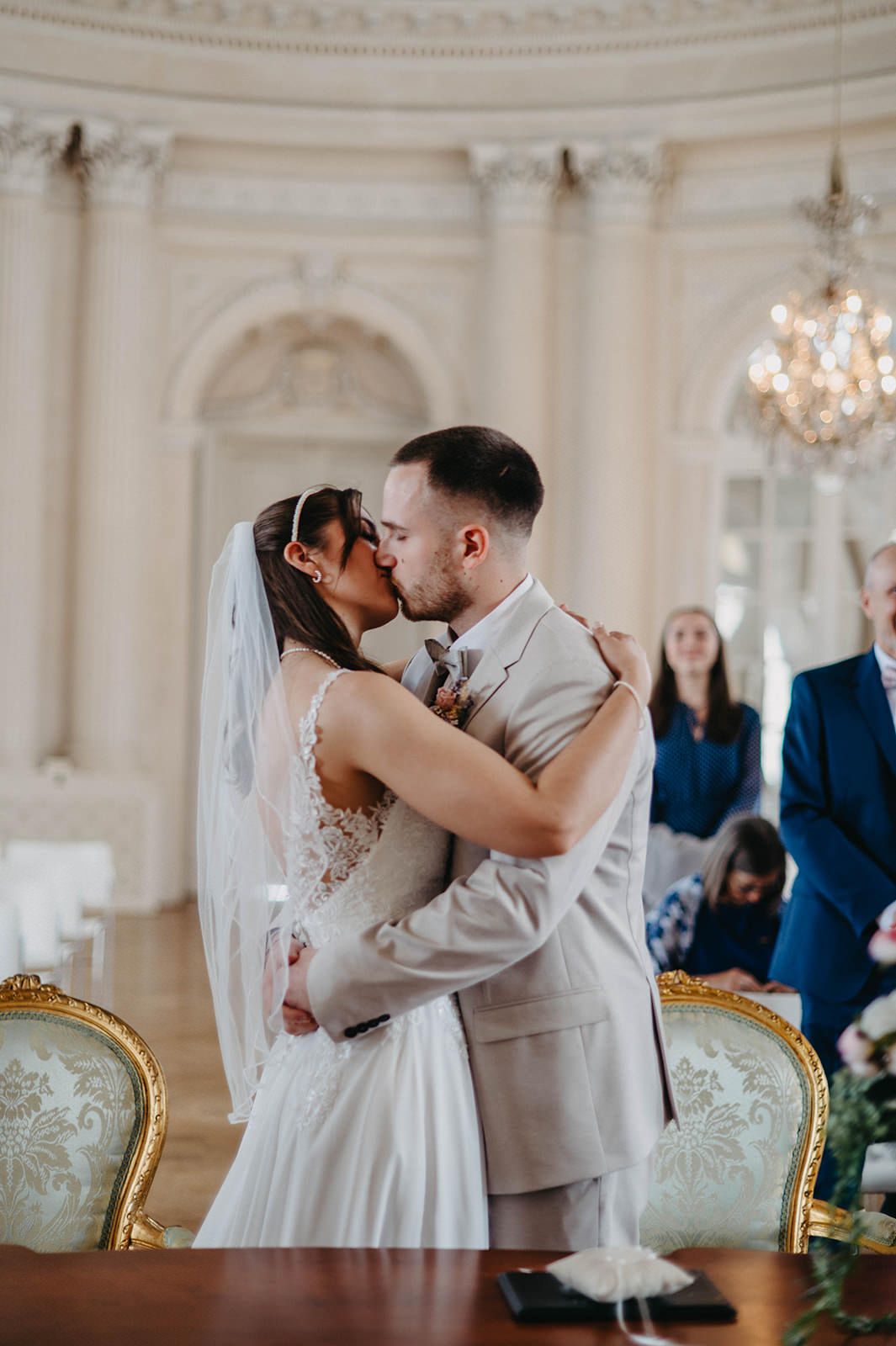 Romantic wedding couple sharing their first kiss in an elegant white ballroom with ornate decor and chandeliers.