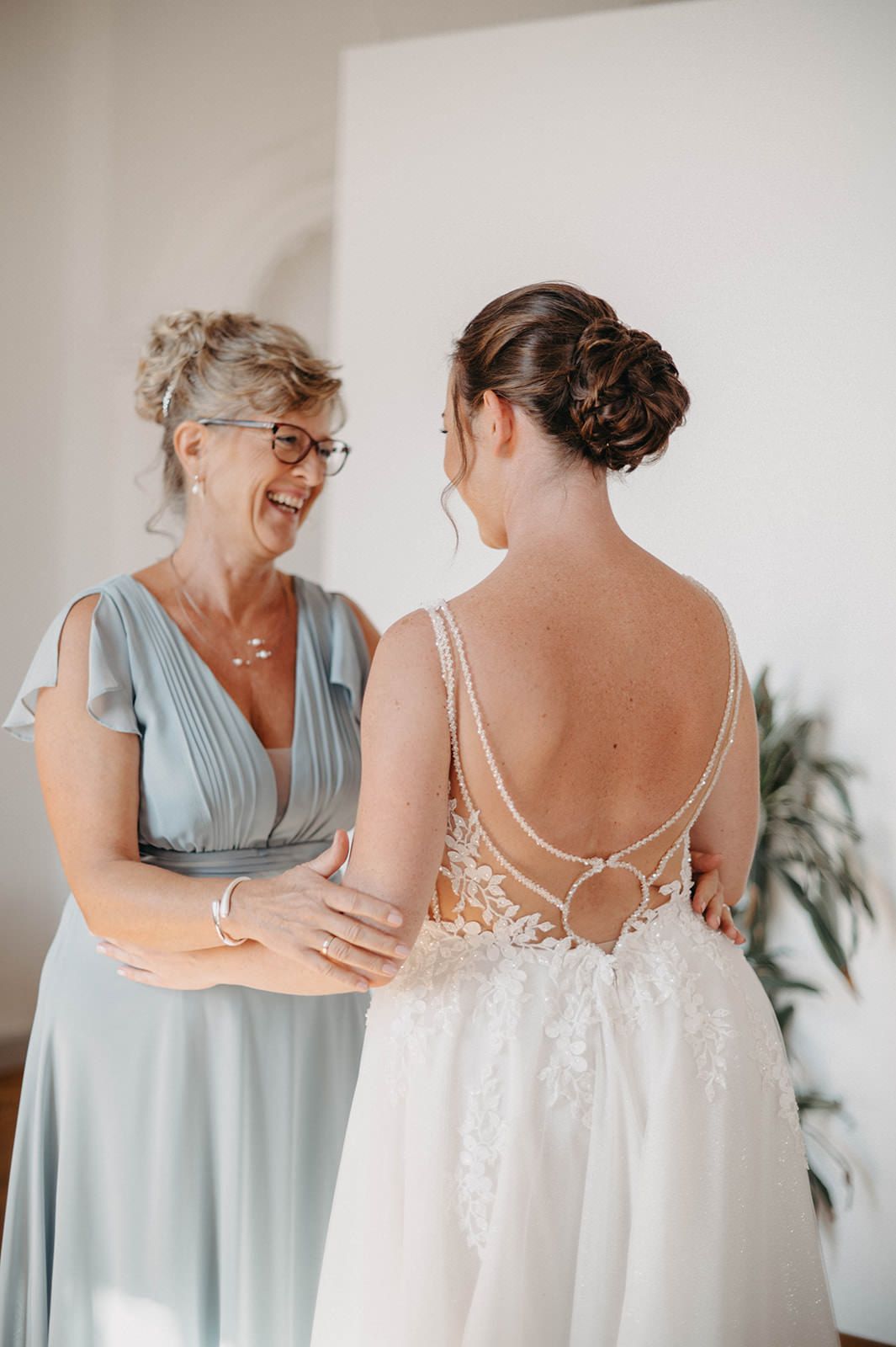 A touching moment between a bride in a white lace gown and someone in a light blue dress during wedding preparations.