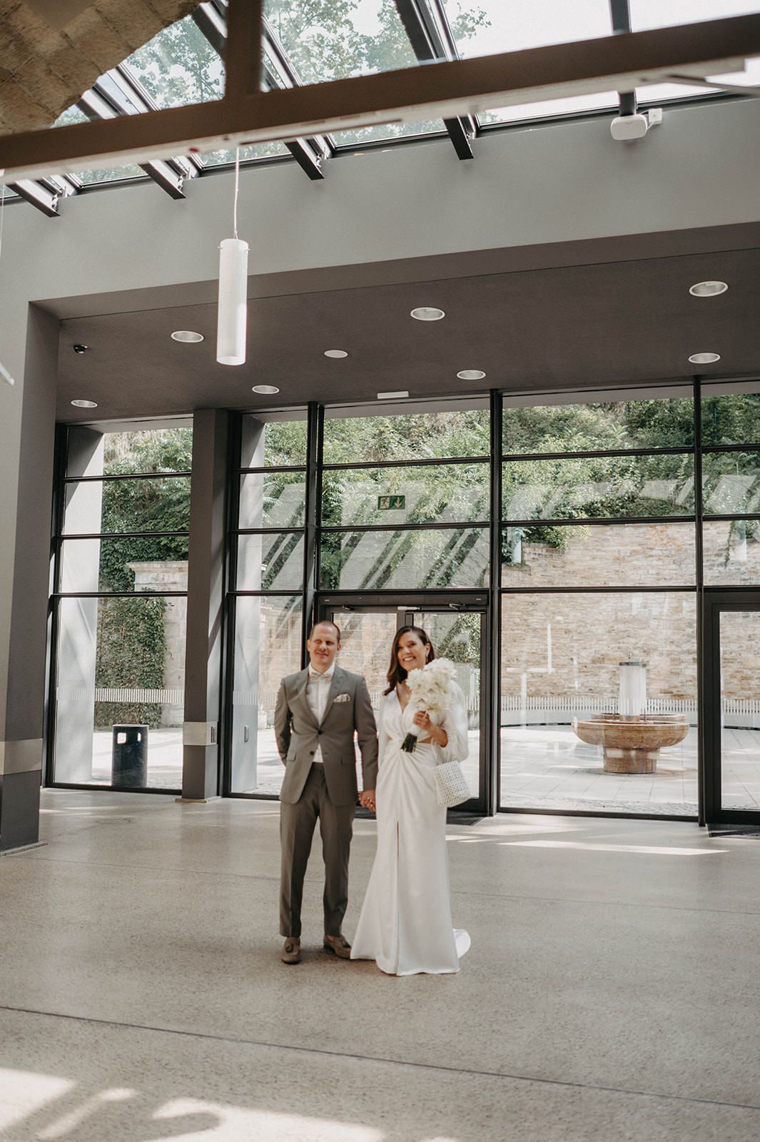 Newlywed couple shares their first dance in a modern event space with floor-to-ceiling windows and exposed beams.