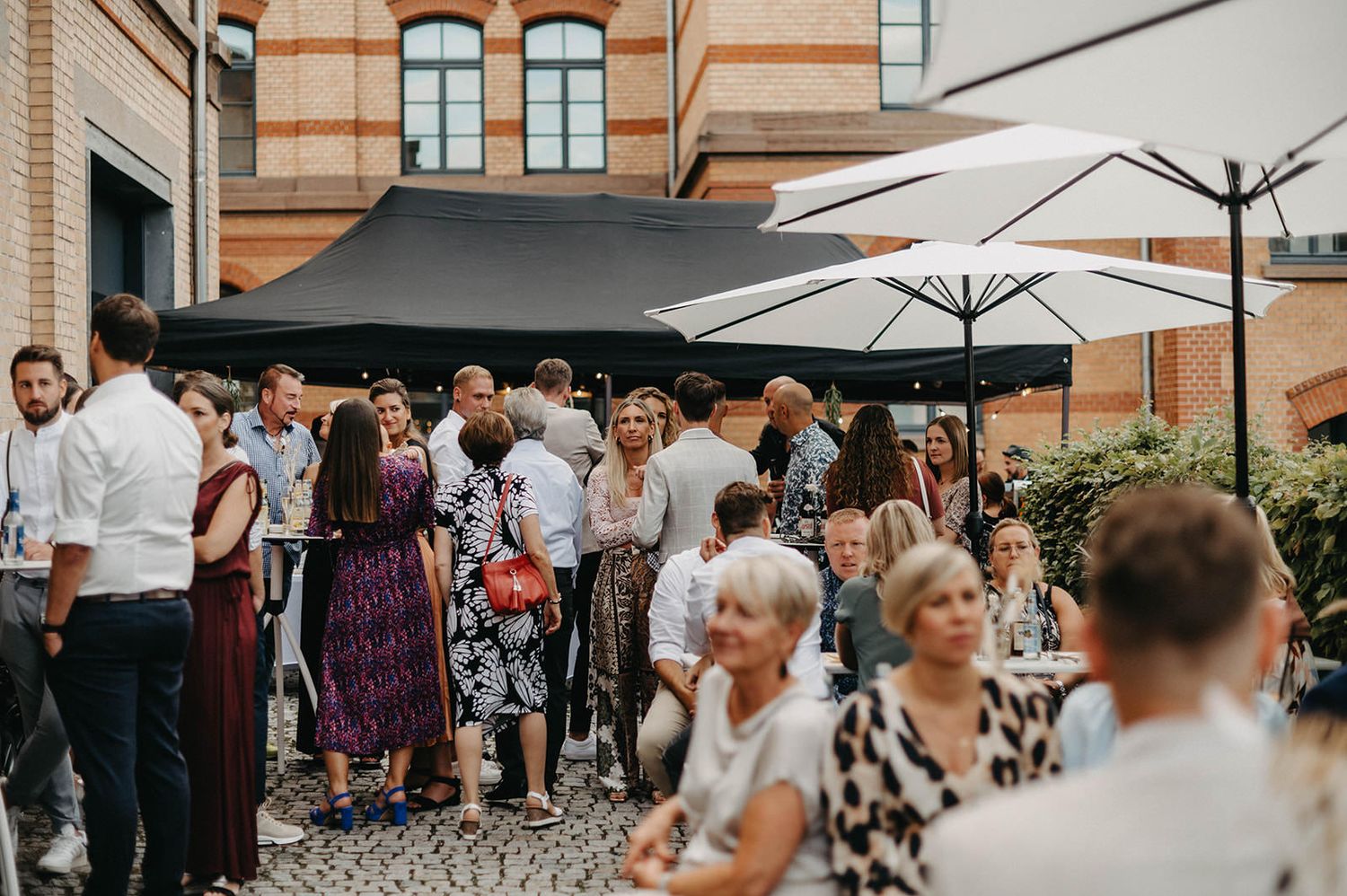 Wedding guests mingle and socialize in an outdoor courtyard with white umbrellas and brick architecture.