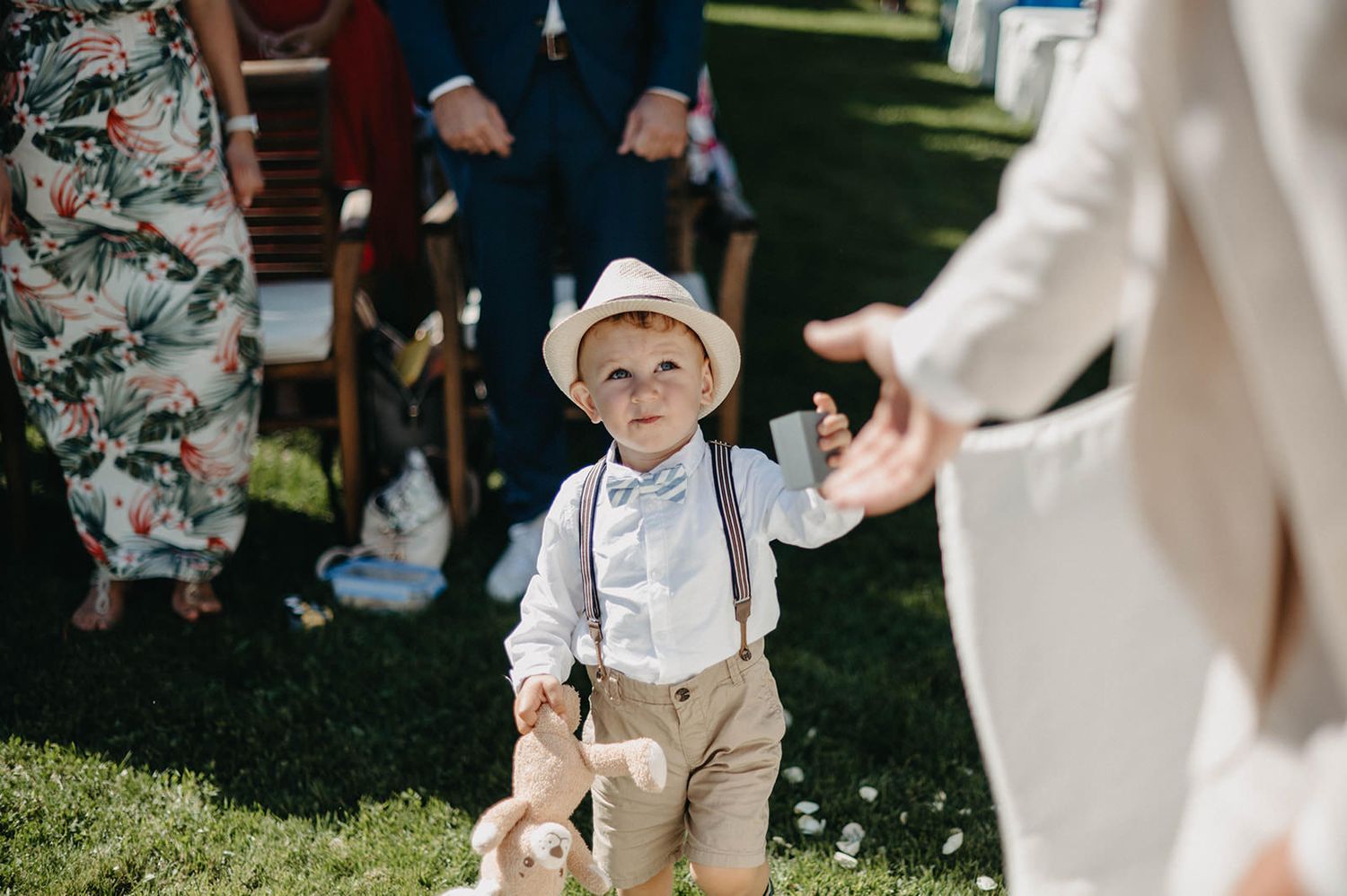 Ring bearer in tan suspenders and white shirt walks down outdoor aisle during wedding ceremony.