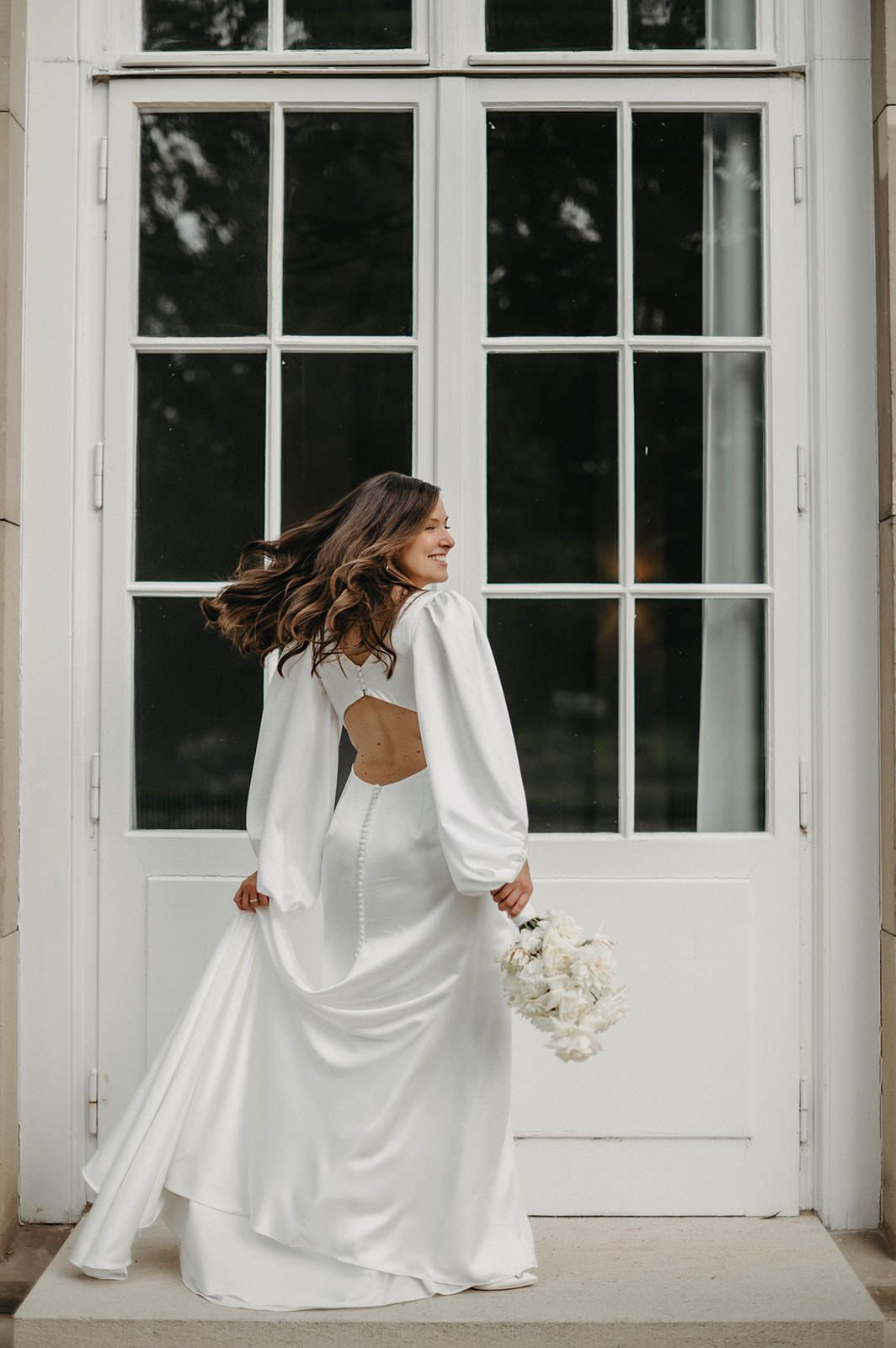 A person in a flowing white wedding dress twirls gracefully on a white porch with large windows in the background.