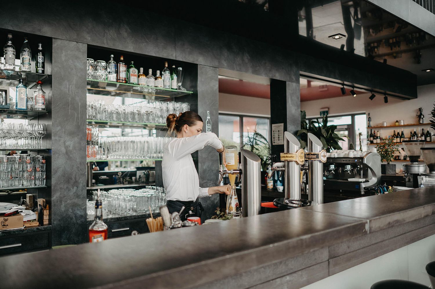 Modern industrial restaurant interior with concrete pillars, long bar counter, and moody atmospheric lighting.