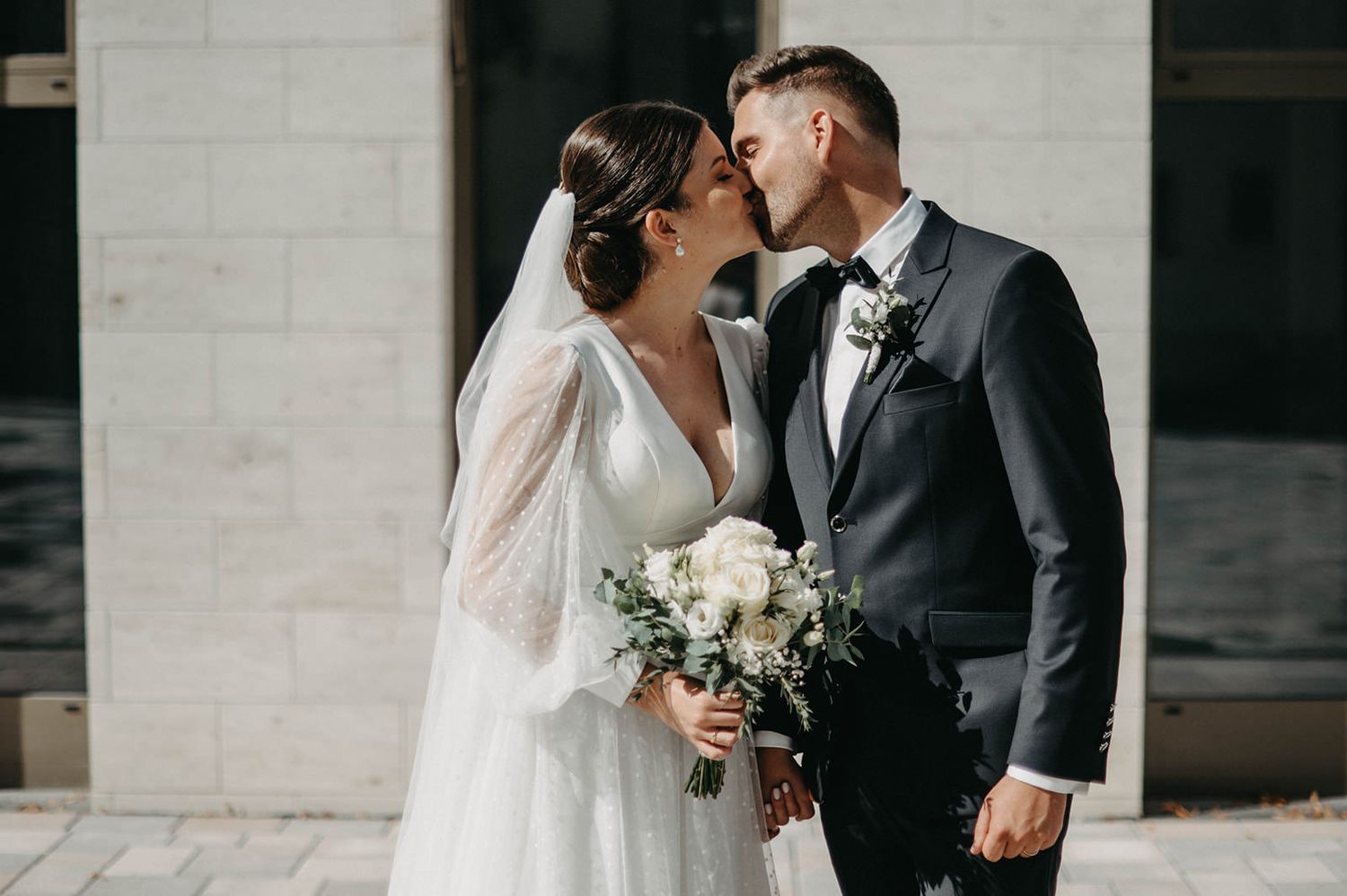 A romantic wedding couple shares an intimate moment outdoors while holding a white floral bouquet.