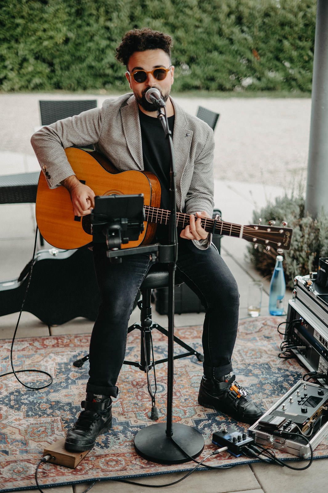 Musician performs with acoustic guitar and microphone setup on patterned rug wearing sunglasses and gray blazer.