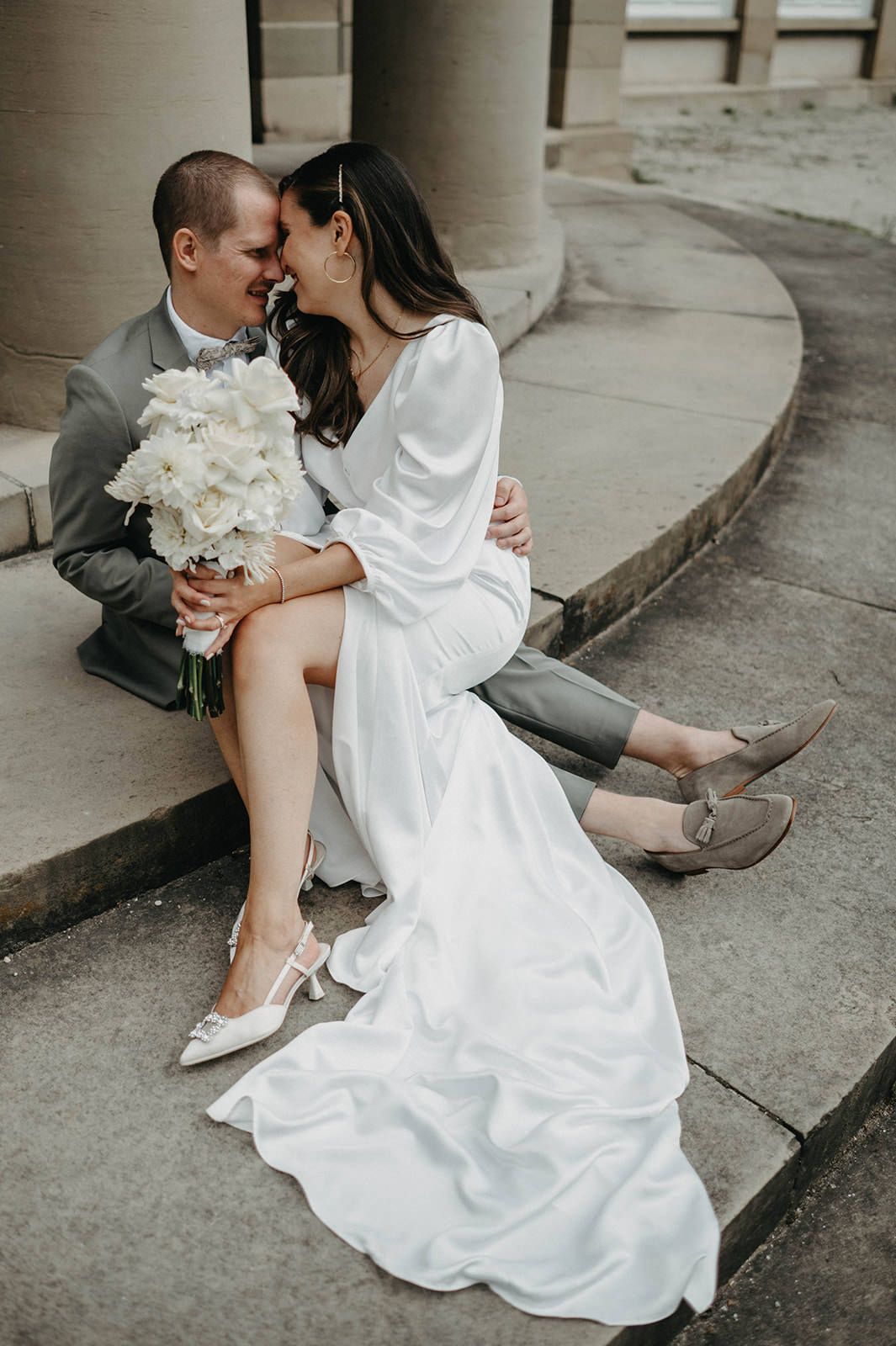 Romantic wedding couple sitting on stone steps with white bouquet and flowing dress in intimate urban setting.