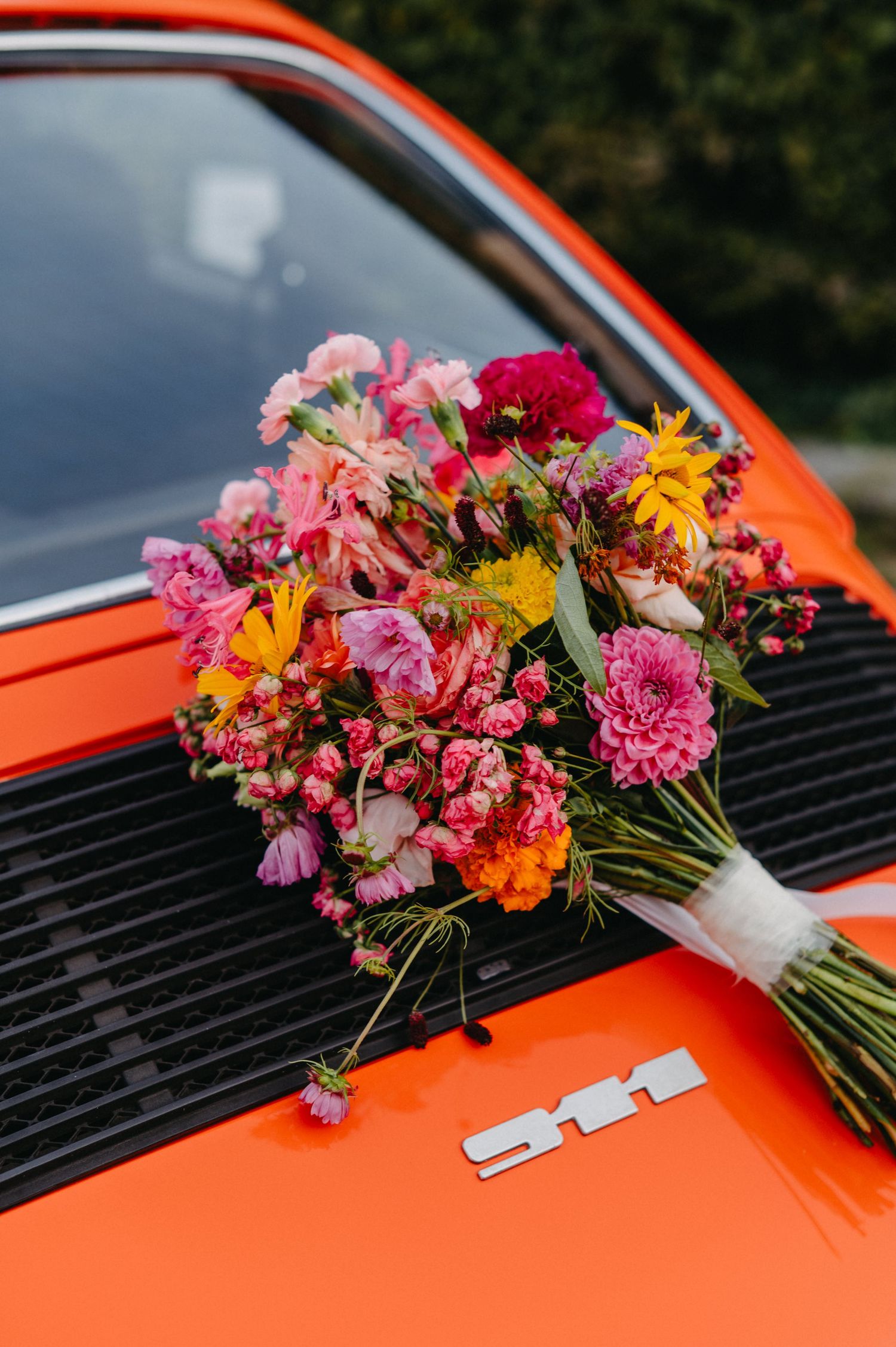 A vibrant bouquet of pink and orange flowers rests on the hood of a bright orange car.