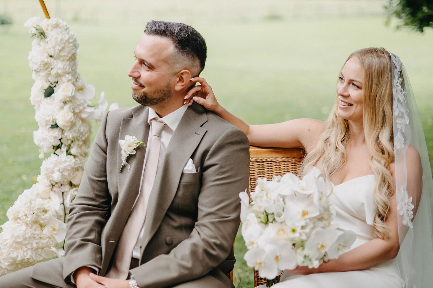 A couple sits together at their outdoor wedding ceremony surrounded by elegant white orchid floral arrangements.