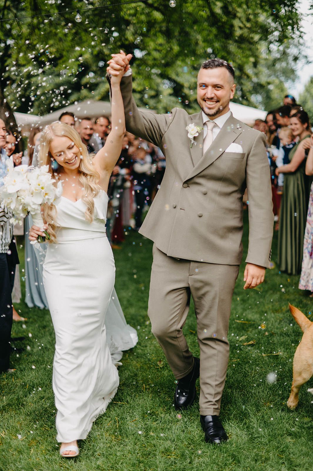 A bride and groom walk together on grass while guests throw confetti during an outdoor wedding celebration.