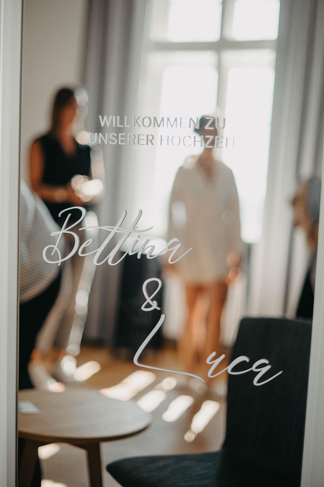 Elegant white wedding welcome sign with calligraphy text displayed in a bright interior venue with window lighting.