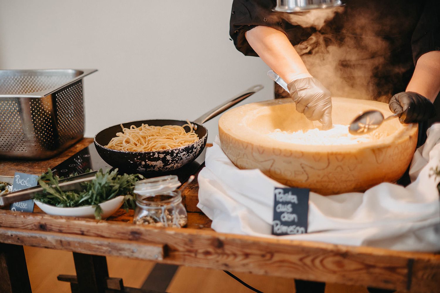 Chef tossing pasta in a large wheel of cheese with fresh ingredients and skillets on wooden table.