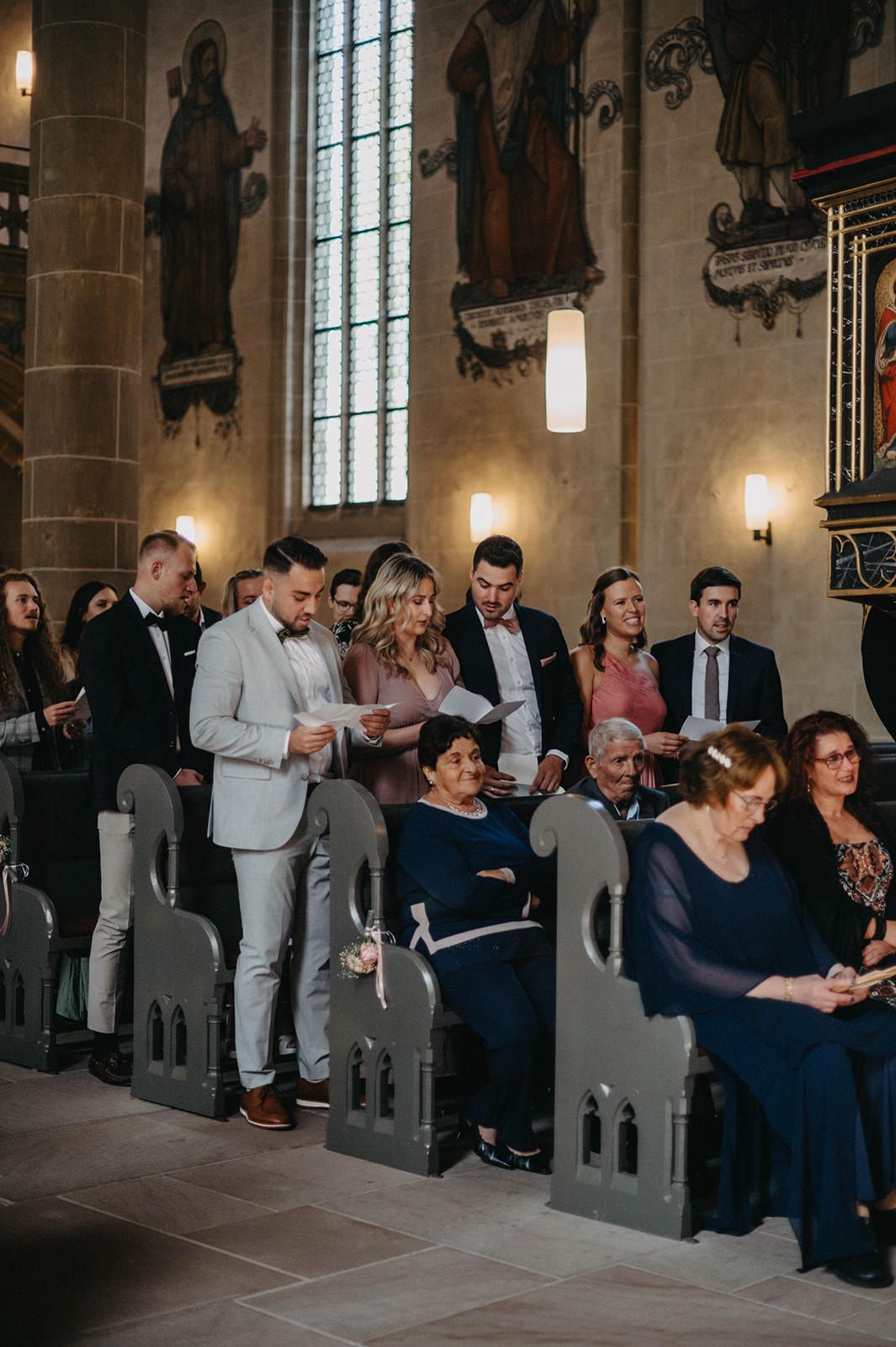 Wedding guests seated in wooden church pews with religious artwork and stained glass windows visible in the background.