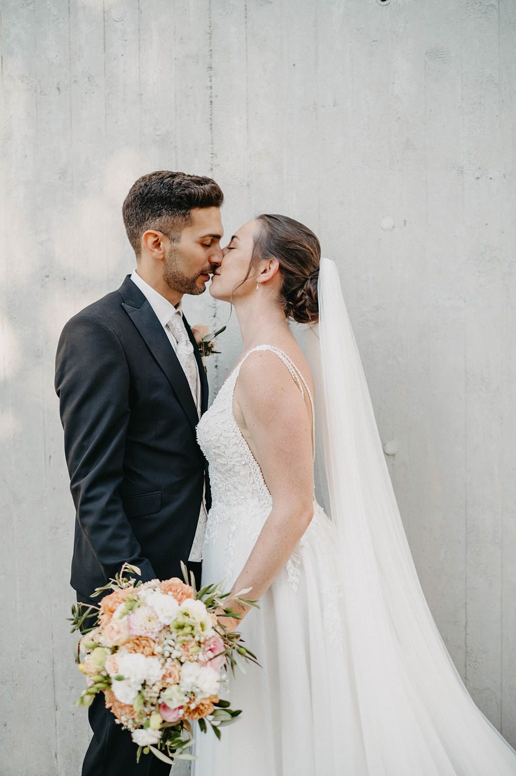 A romantic wedding portrait showing a couple kissing against a gray wall with the bride holding a pink and white bouquet.