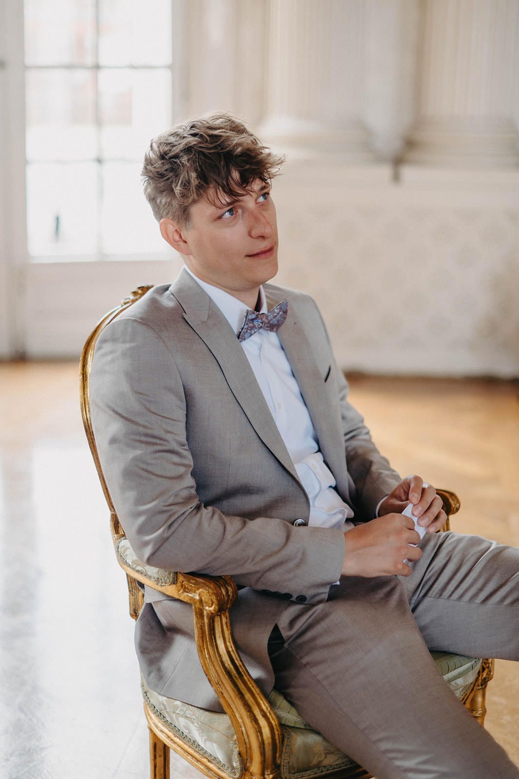 A person in a light gray suit sits casually in an ornate gold chair against a bright white interior.