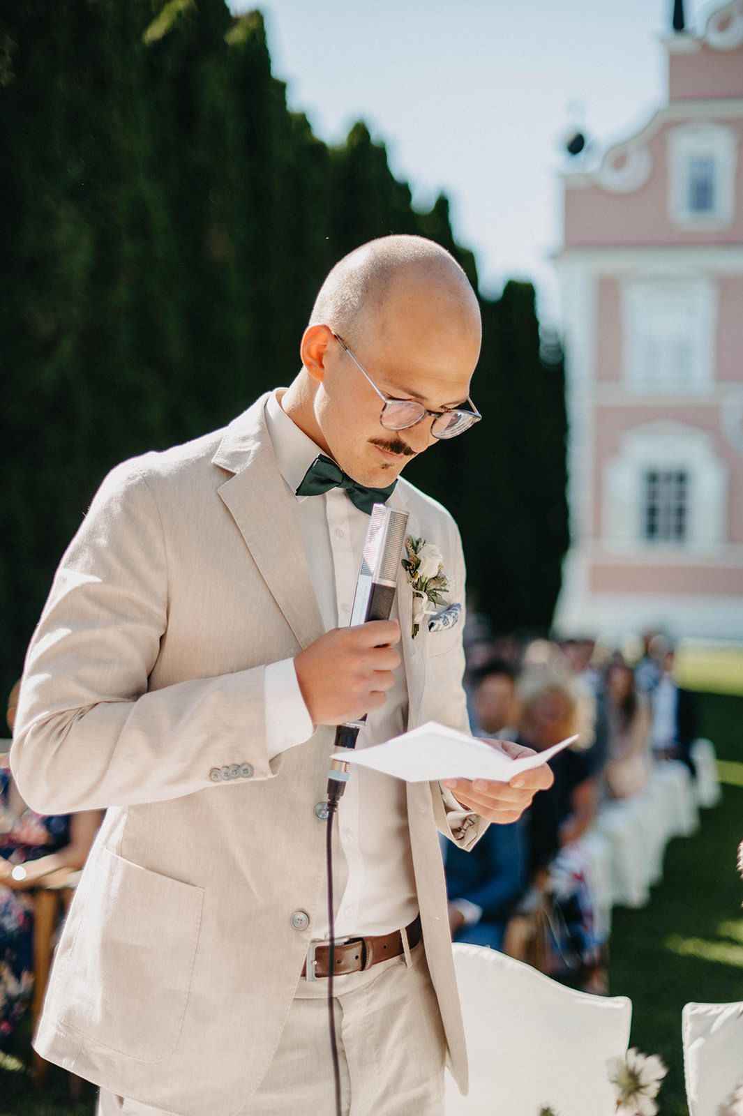 Emotional groom in a light beige suit wipes away tears during an outdoor wedding ceremony.